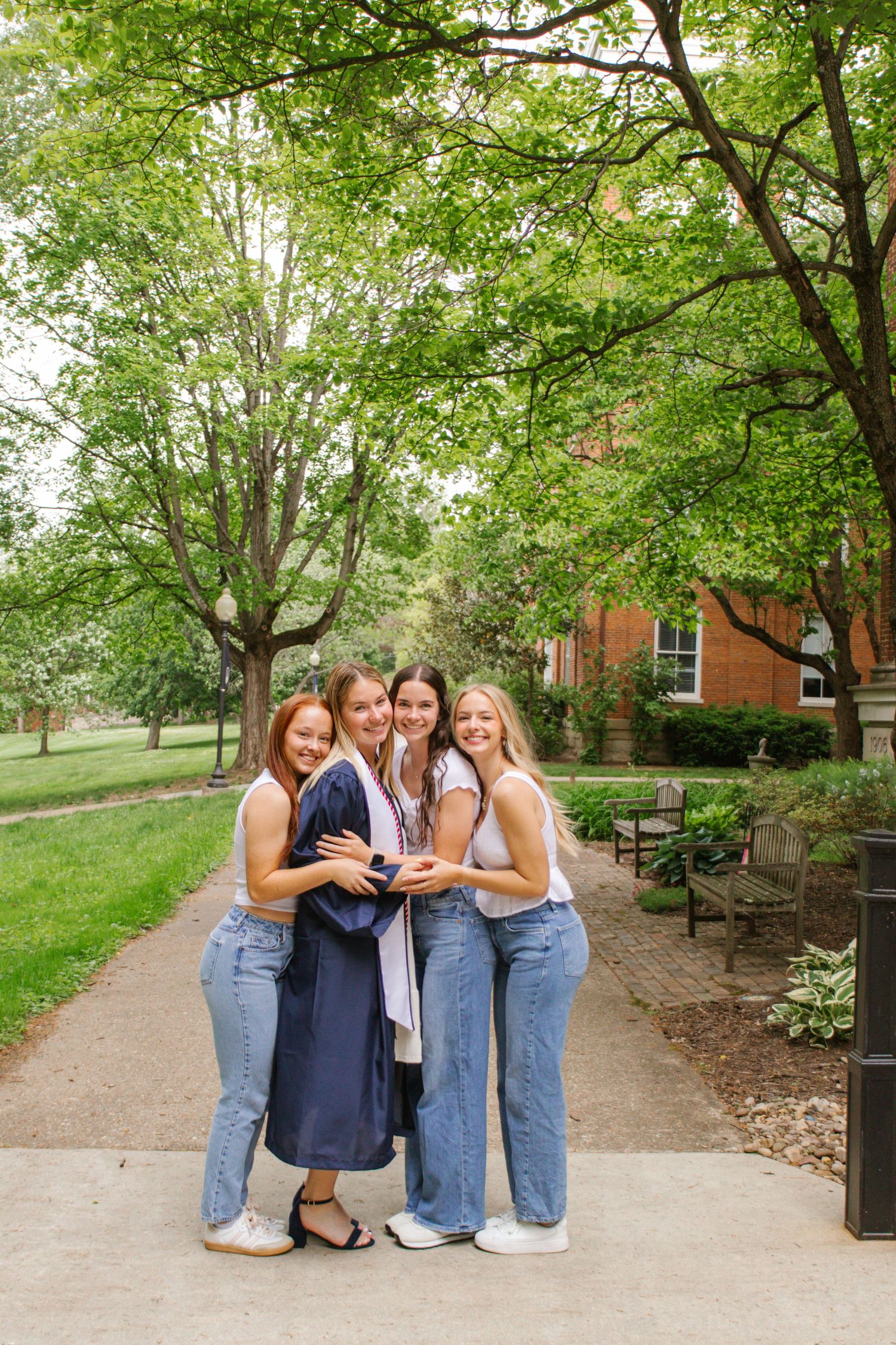 Four young women in jeans hugging and smiling outdoors on a college campus pathway, one wearing a graduation gown.