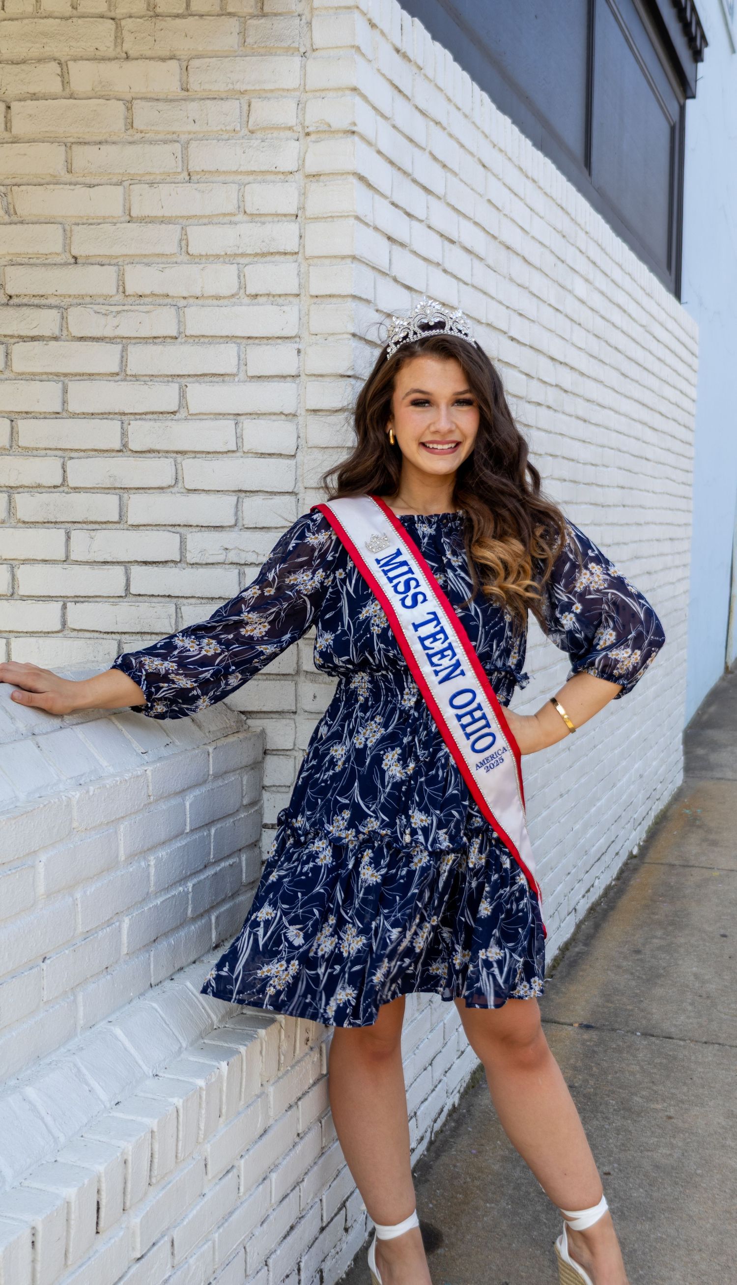 Miss Teen Ohio beauty queen smiles in navy floral dress with crown and sash by brick wall.