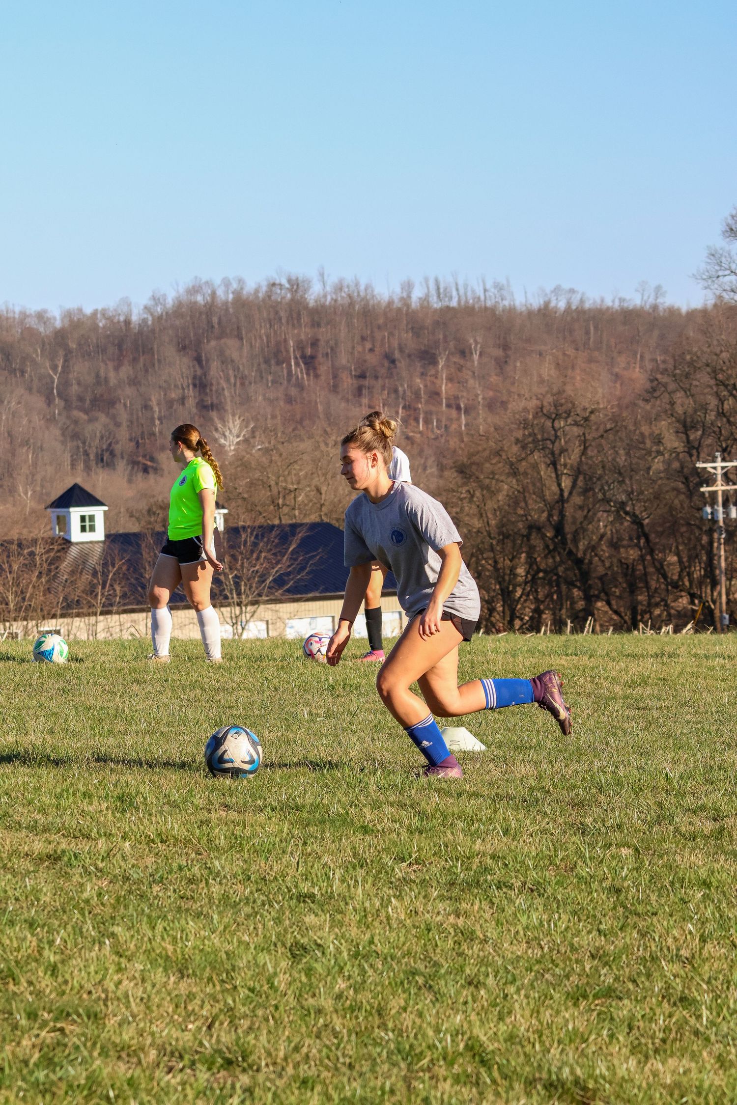 Female soccer player in gray jersey dribbles ball on green grass field during practice.