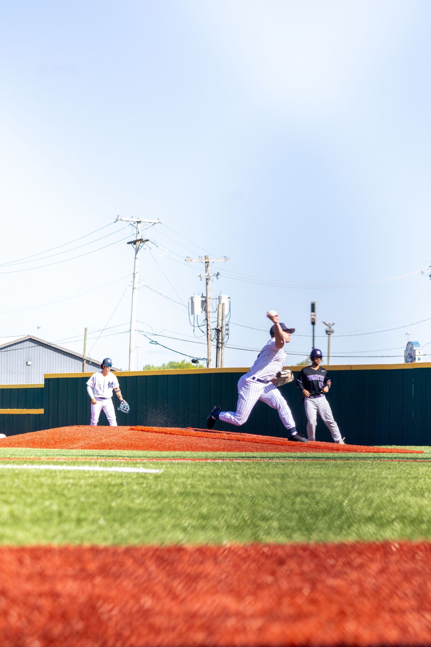 A baseball pitcher winds up to throw on a red dirt mound during a sunny outdoor game with players and green outfield wall behind.