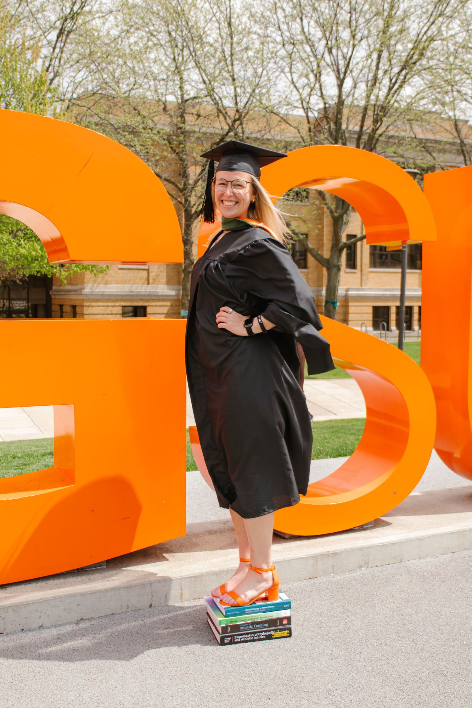 Female graduate in black cap and gown stands on books beside large orange BGSU letters on campus.