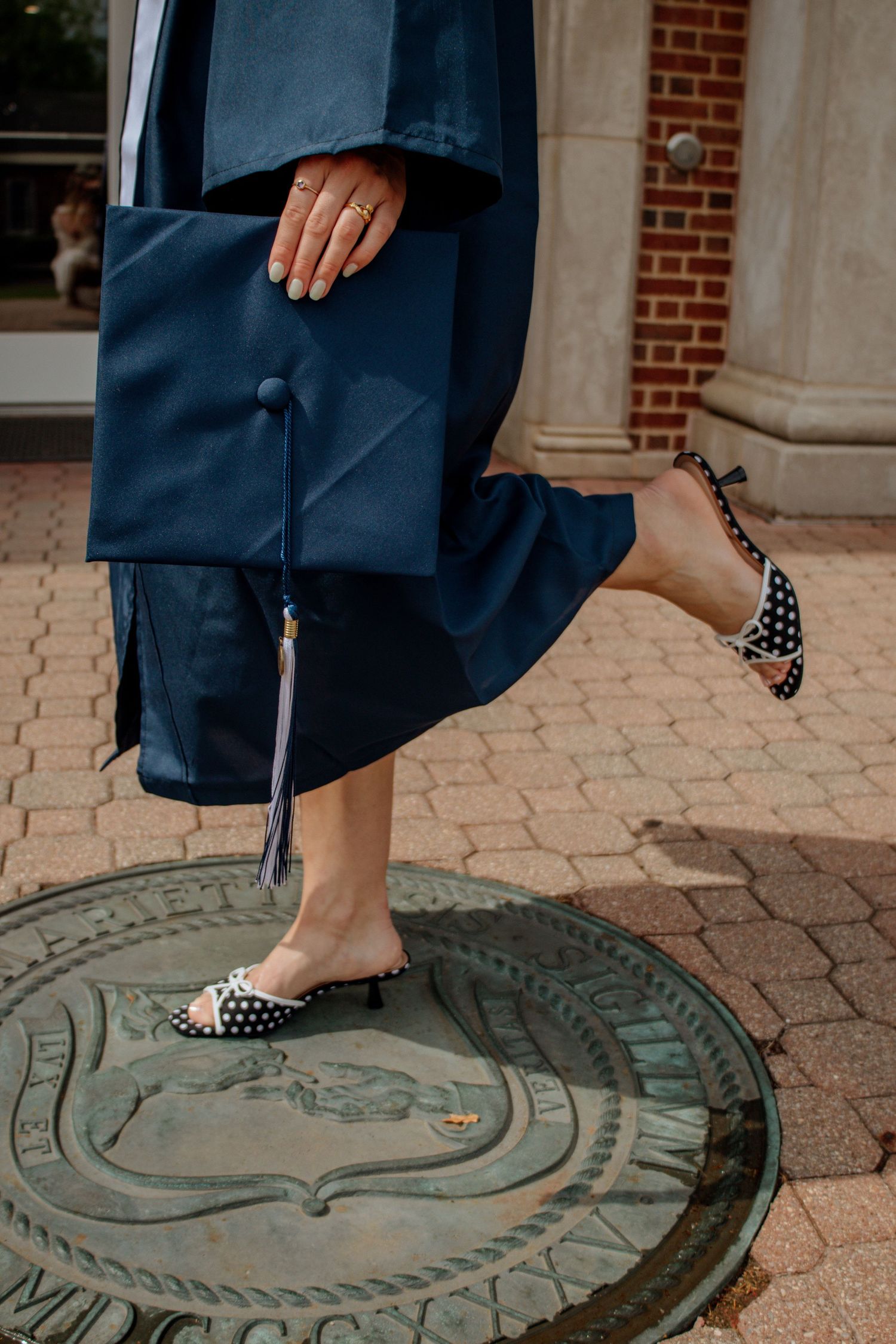 Close-up of graduate in navy gown standing on bronze university seal wearing polka dot heels holding cap.