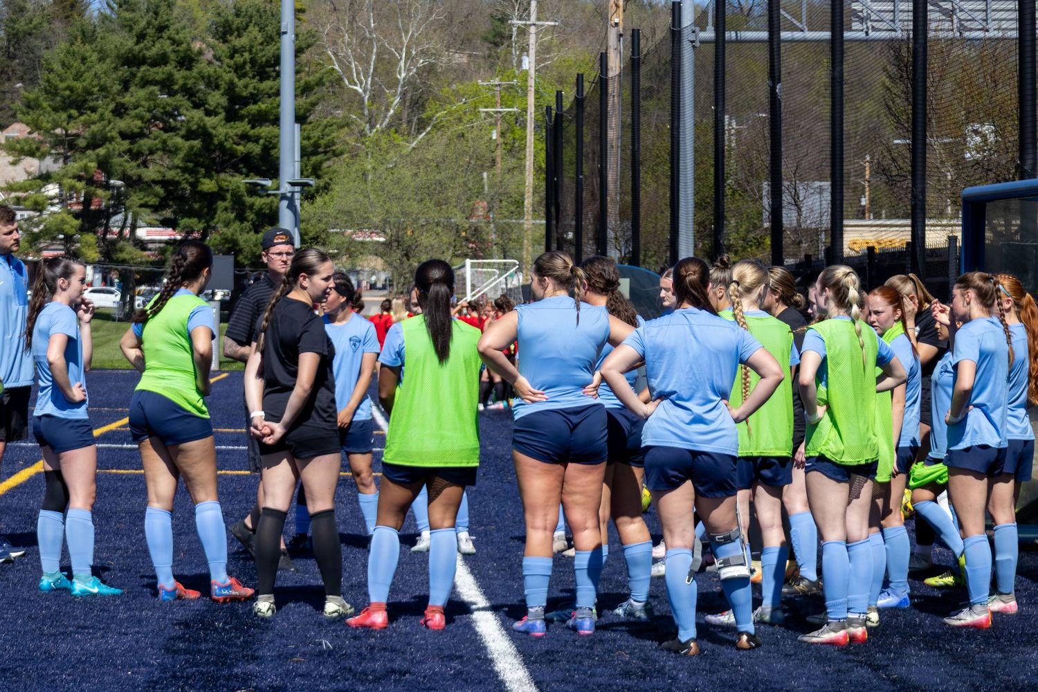 Womens soccer team huddles together on blue turf field during outdoor practice session.