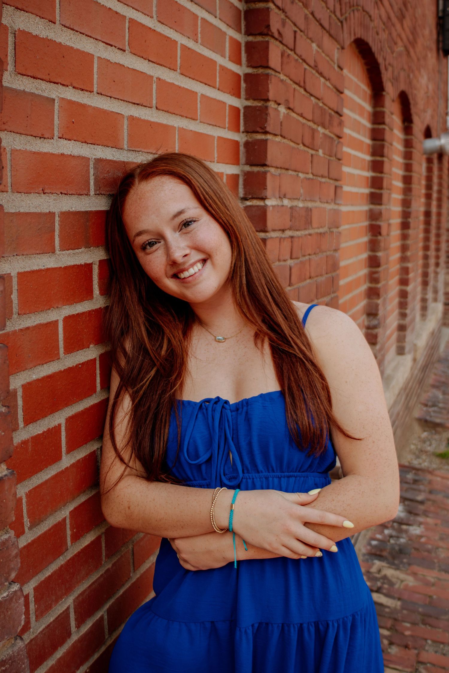 Smiling redhead woman in a royal blue dress leaning against a brick wall.