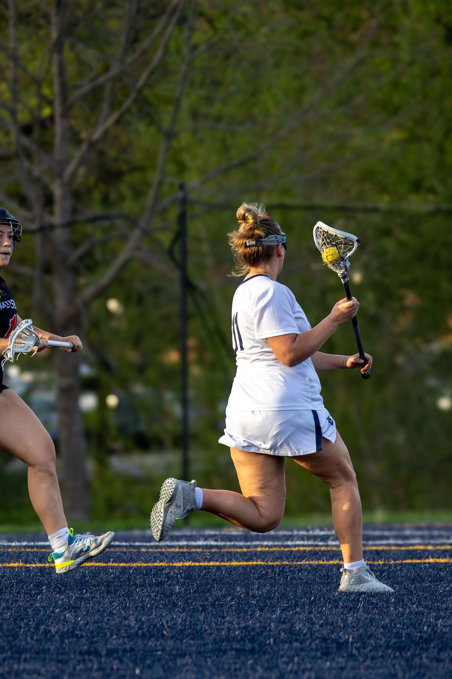 Female lacrosse player in white uniform runs with stick during outdoor game on blue turf field.