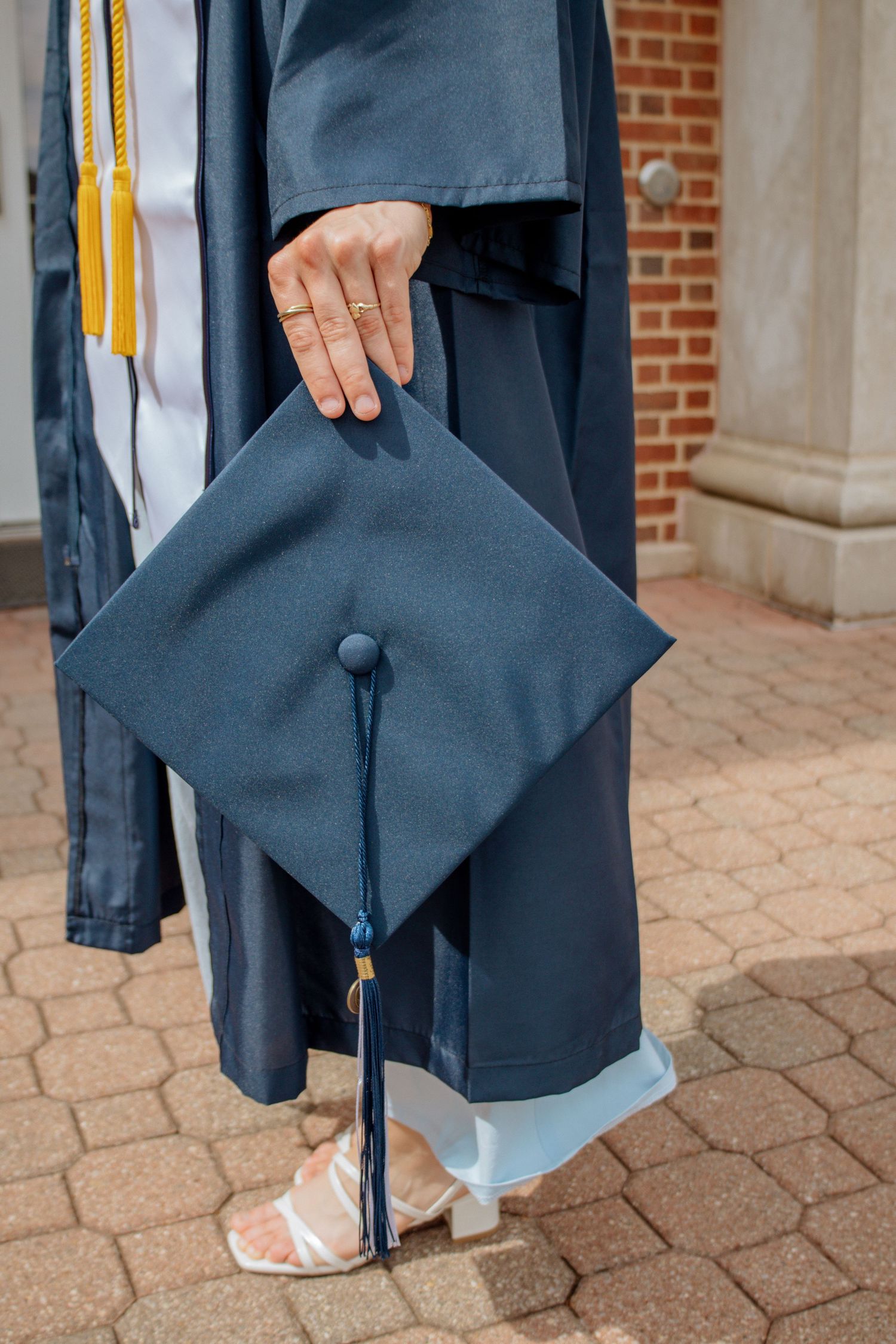 Close-up of graduate in navy gown holding mortarboard cap with blue tassel on brick campus pathway.
