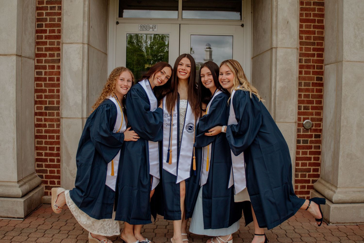 Five college graduates in navy blue robes pose joyfully together outside a brick building on graduation day.