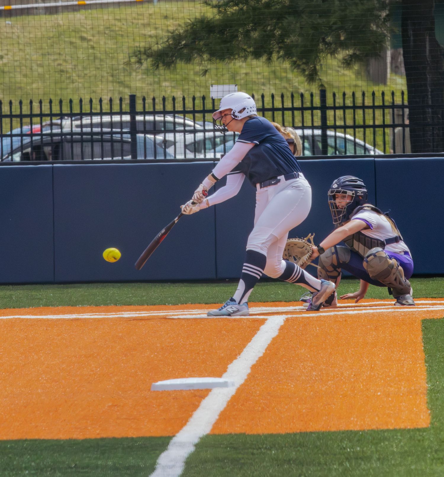 Softball batter in navy uniform swings at yellow ball while catcher crouches behind home plate on orange turf field.