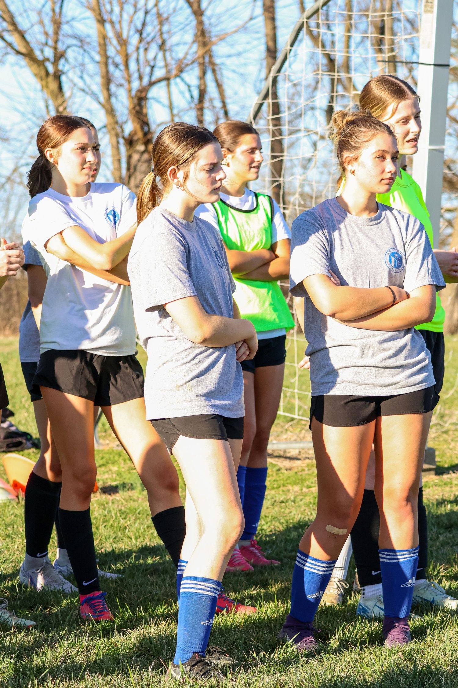 Female soccer players in gray and green jerseys stand near goal during team practice.