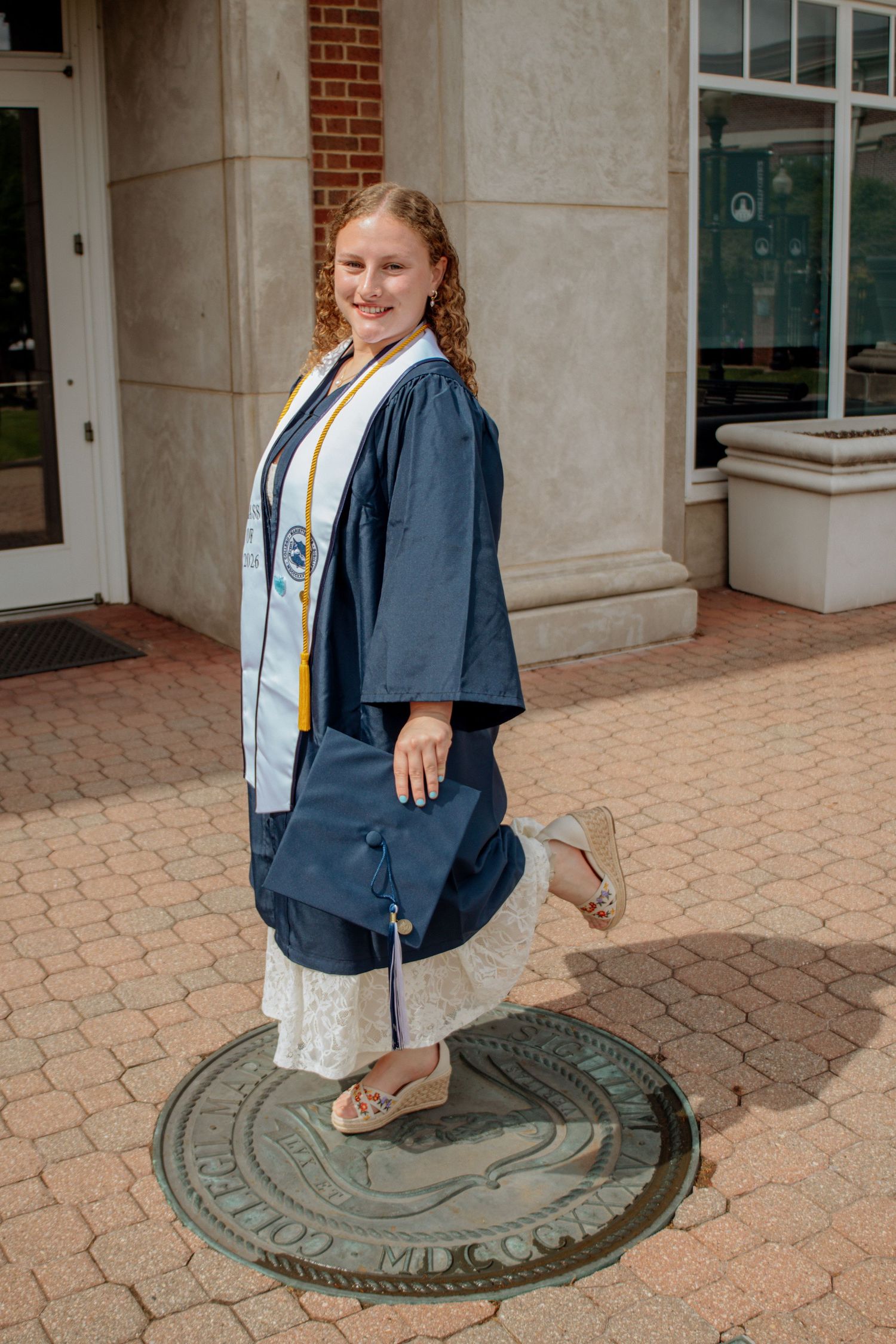 Smiling blonde graduate in navy gown with gold and white stoles poses on university seal holding cap.