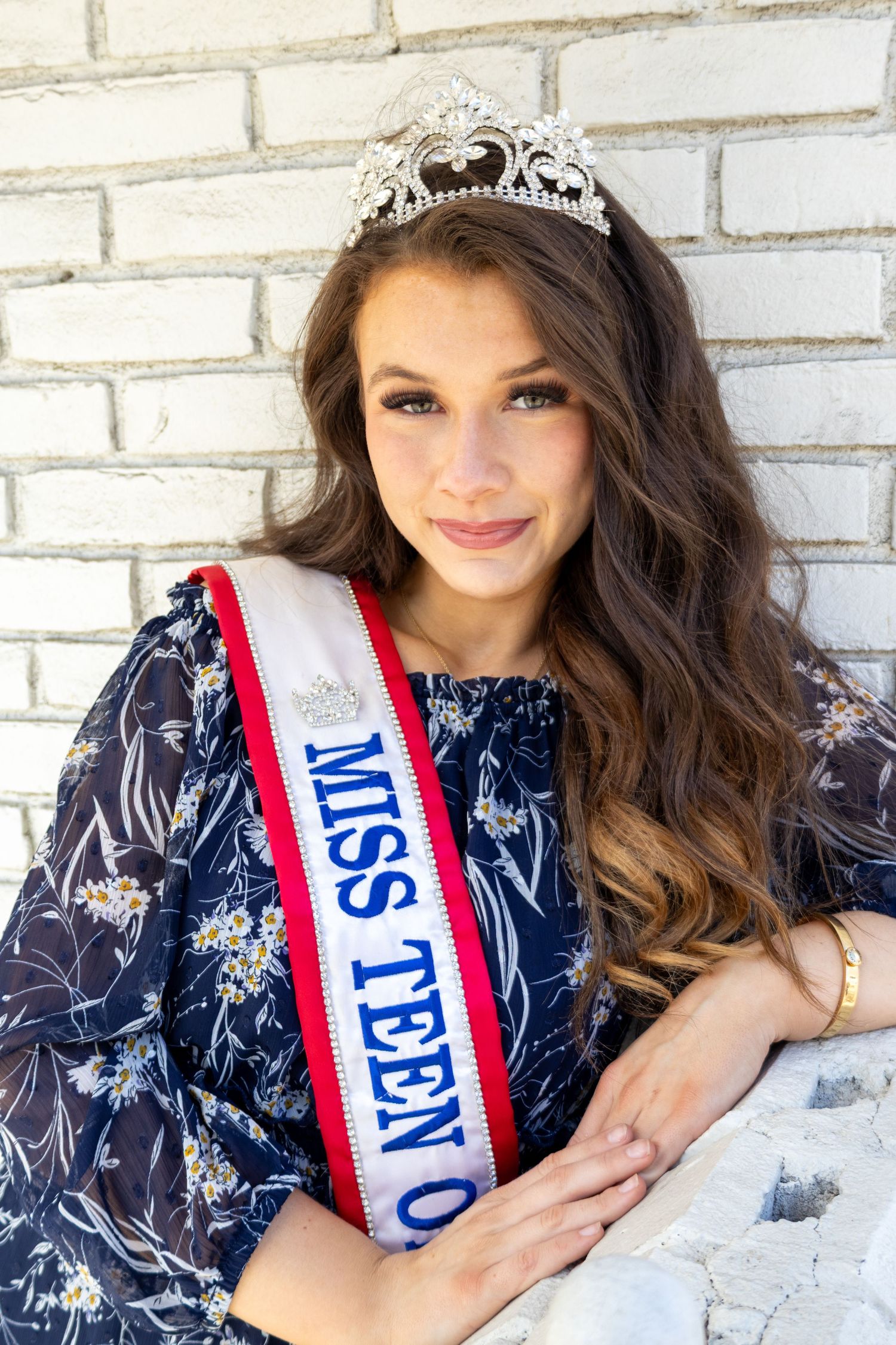 Miss Teen Ohio titleholder in navy floral dress and diamond crown poses against white brick wall.