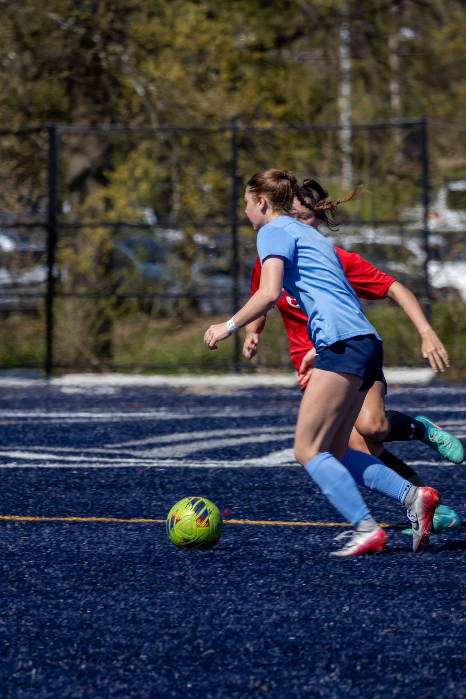 Two girls compete for a soccer ball during an outdoor match, one in light blue and one in red, on a blue turf field.
