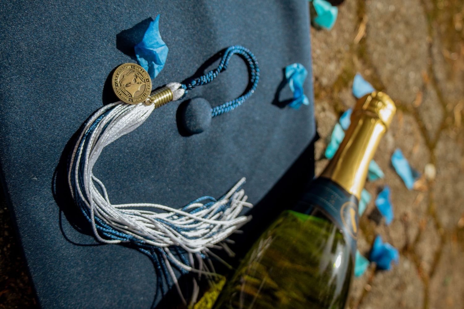 Graduation cap with blue tassel and gold medallion beside a champagne bottle with blue confetti.