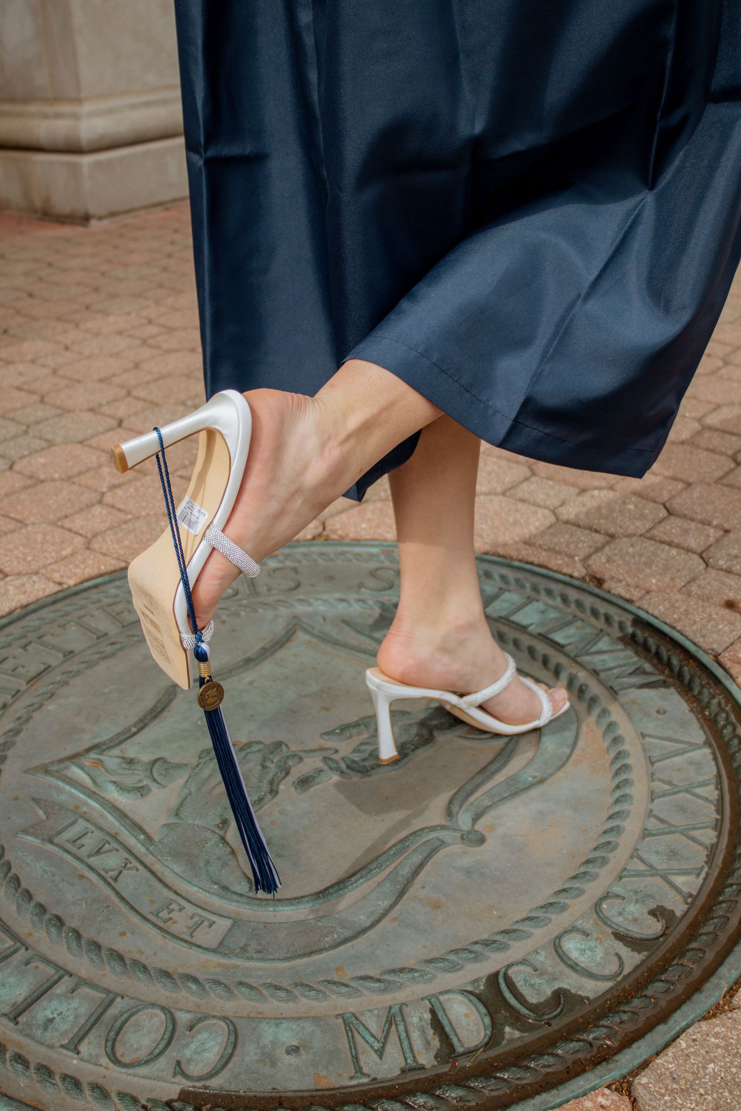 Close-up of graduate feet in white heels and nude sandals standing on bronze university seal medallion.