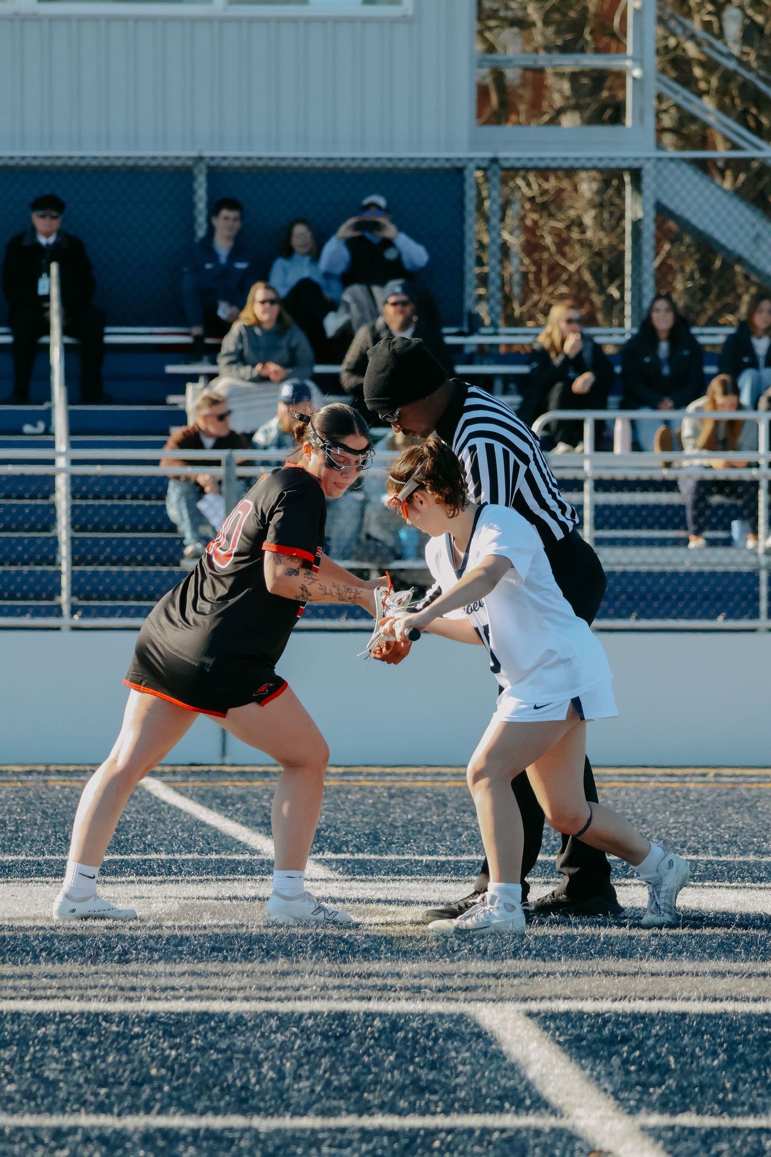 Two female lacrosse players face off with a referee during an outdoor college game.