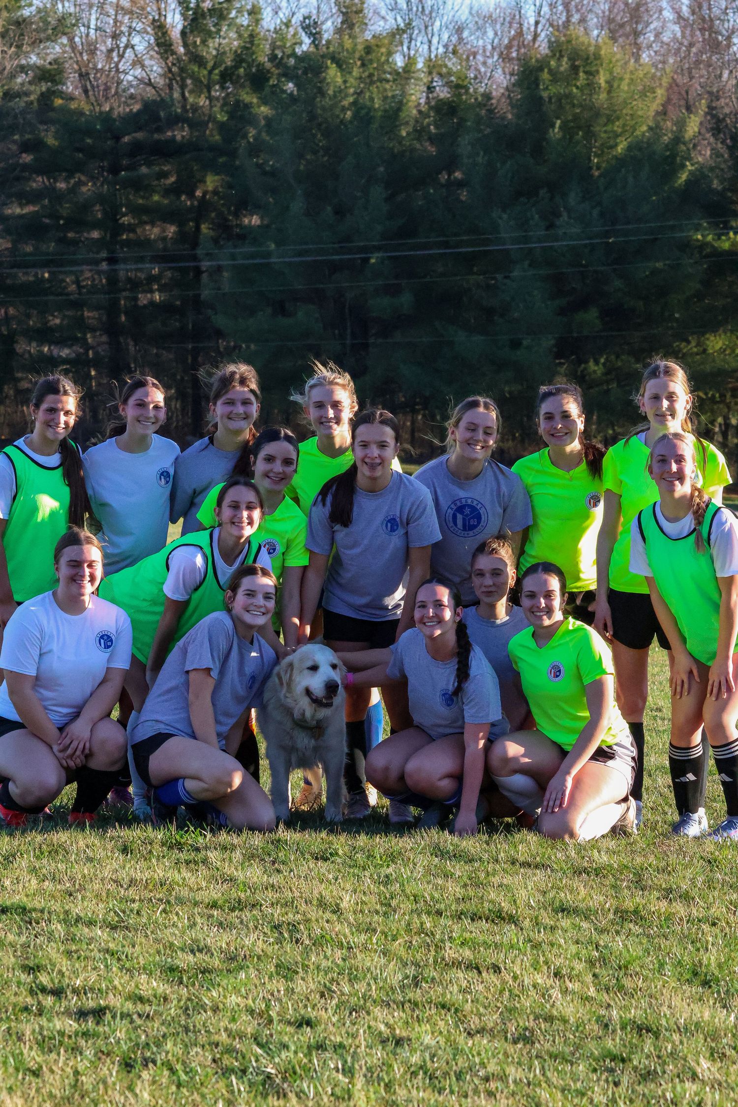 Girls soccer team poses for group photo with golden retriever dog on grass field.