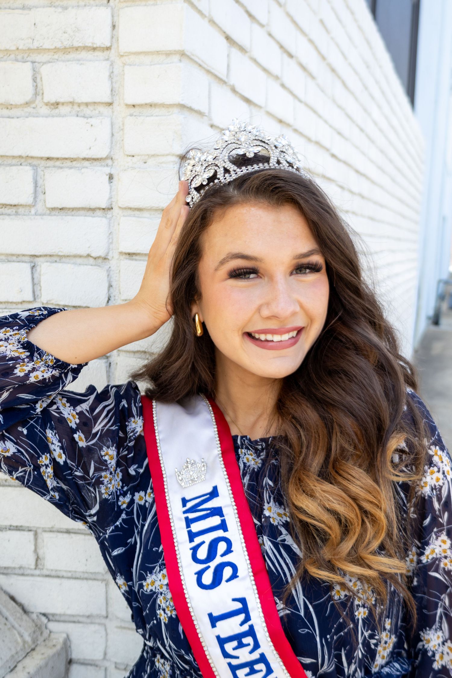 Smiling Miss Teen beauty queen adjusts diamond crown wearing navy floral dress and red sash.