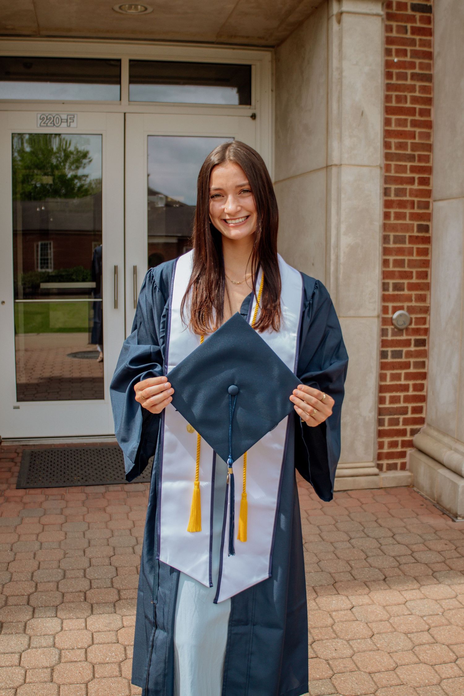 Brunette graduate in navy gown holds cap with gold tassels while smiling outside brick university building.