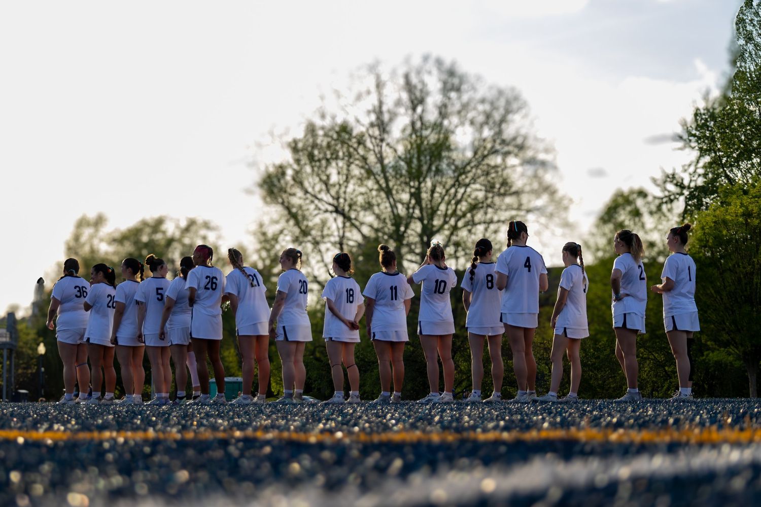 A row of athletes in white uniforms stand together on a track surrounded by green trees at dusk.
