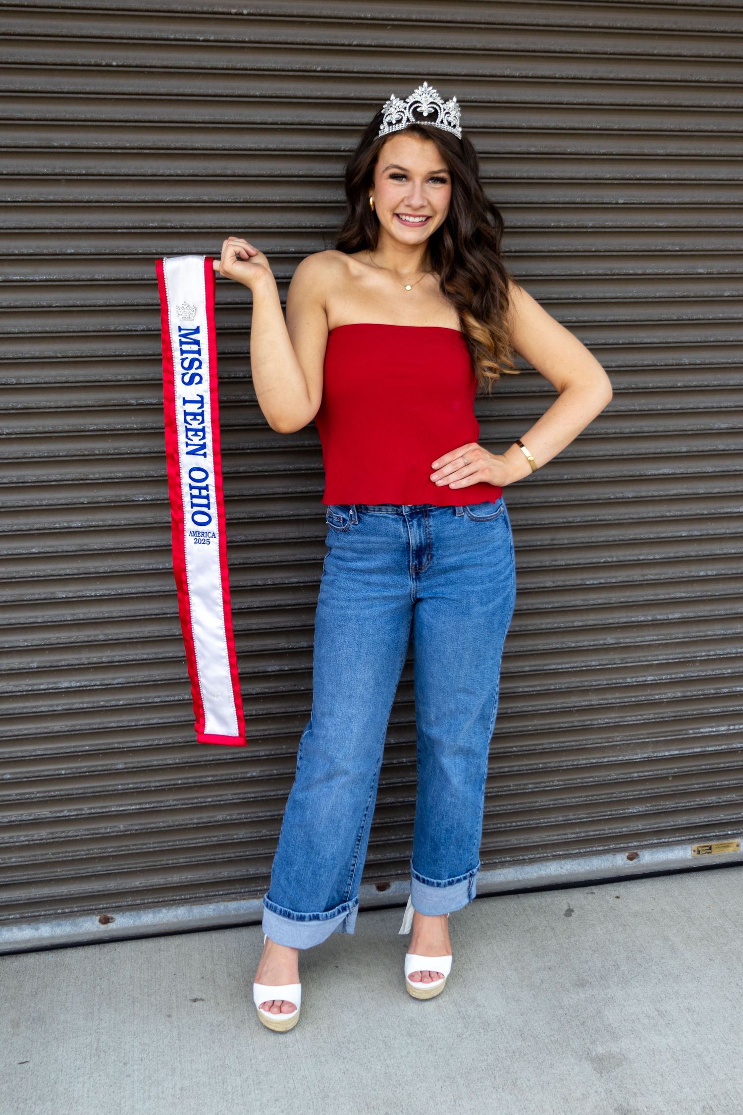 Young woman in red top and jeans holding Miss Teen Ohio sash and wearing crown poses against metal wall.