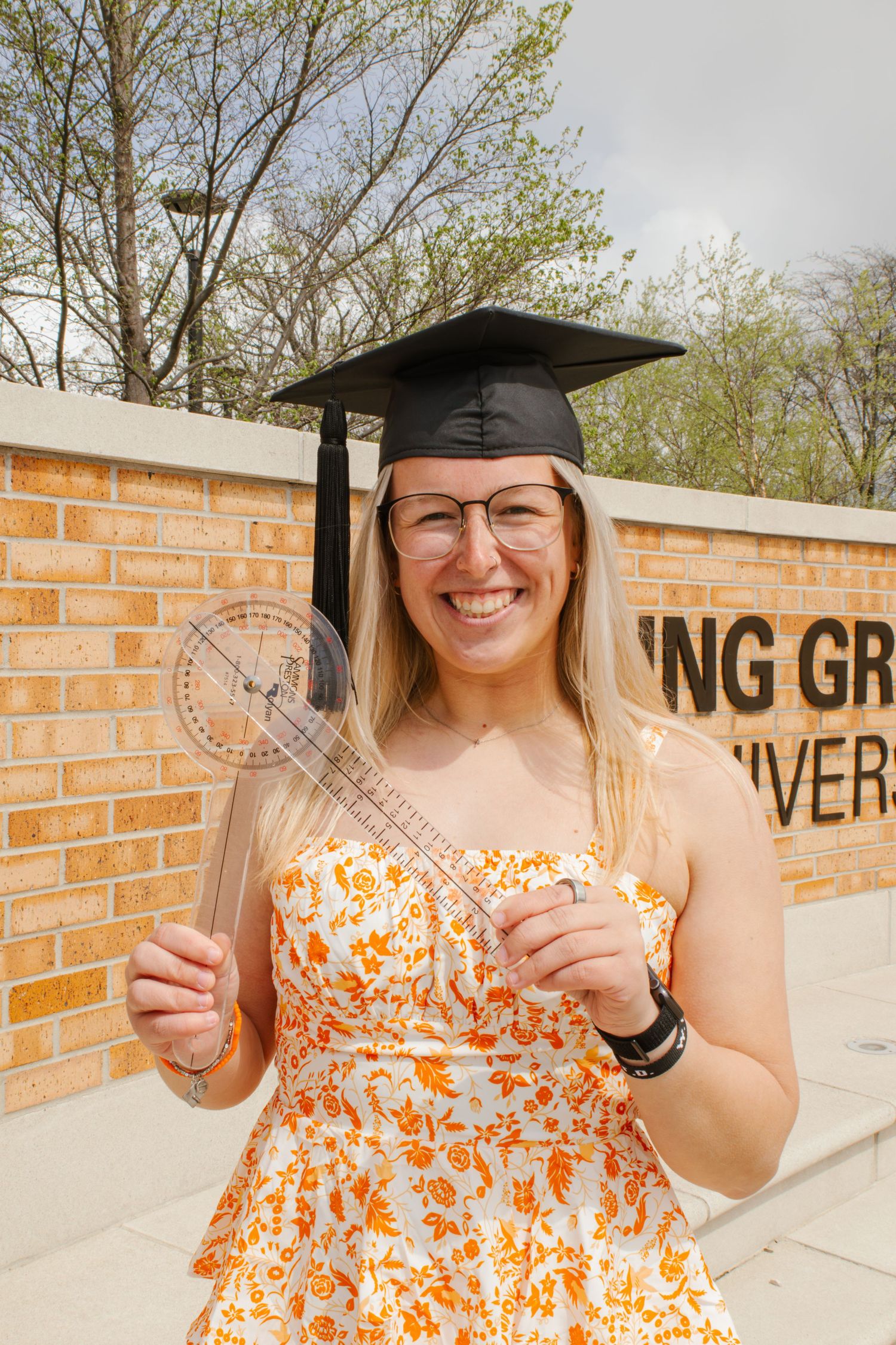 Smiling female graduate in black mortarboard holds a protractor outside Bowling Green University wall.