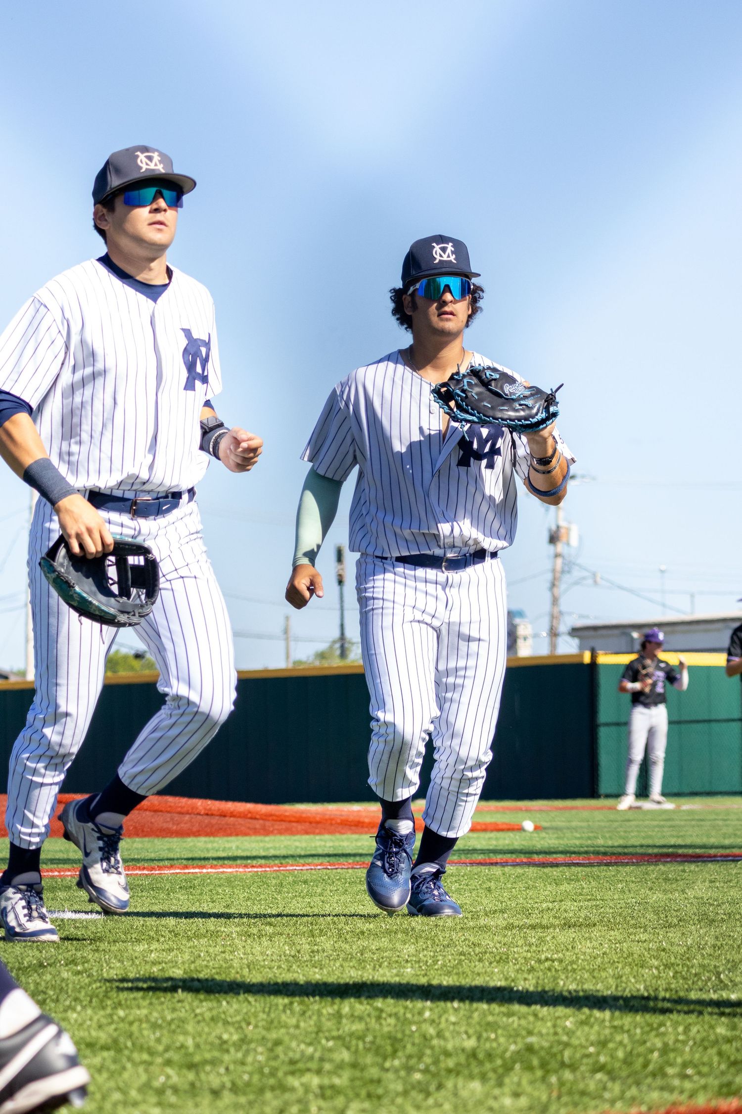 Two baseball players in white pinstripe uniforms jogging on a sunny field, one holding a glove, with a teammate visible in background.