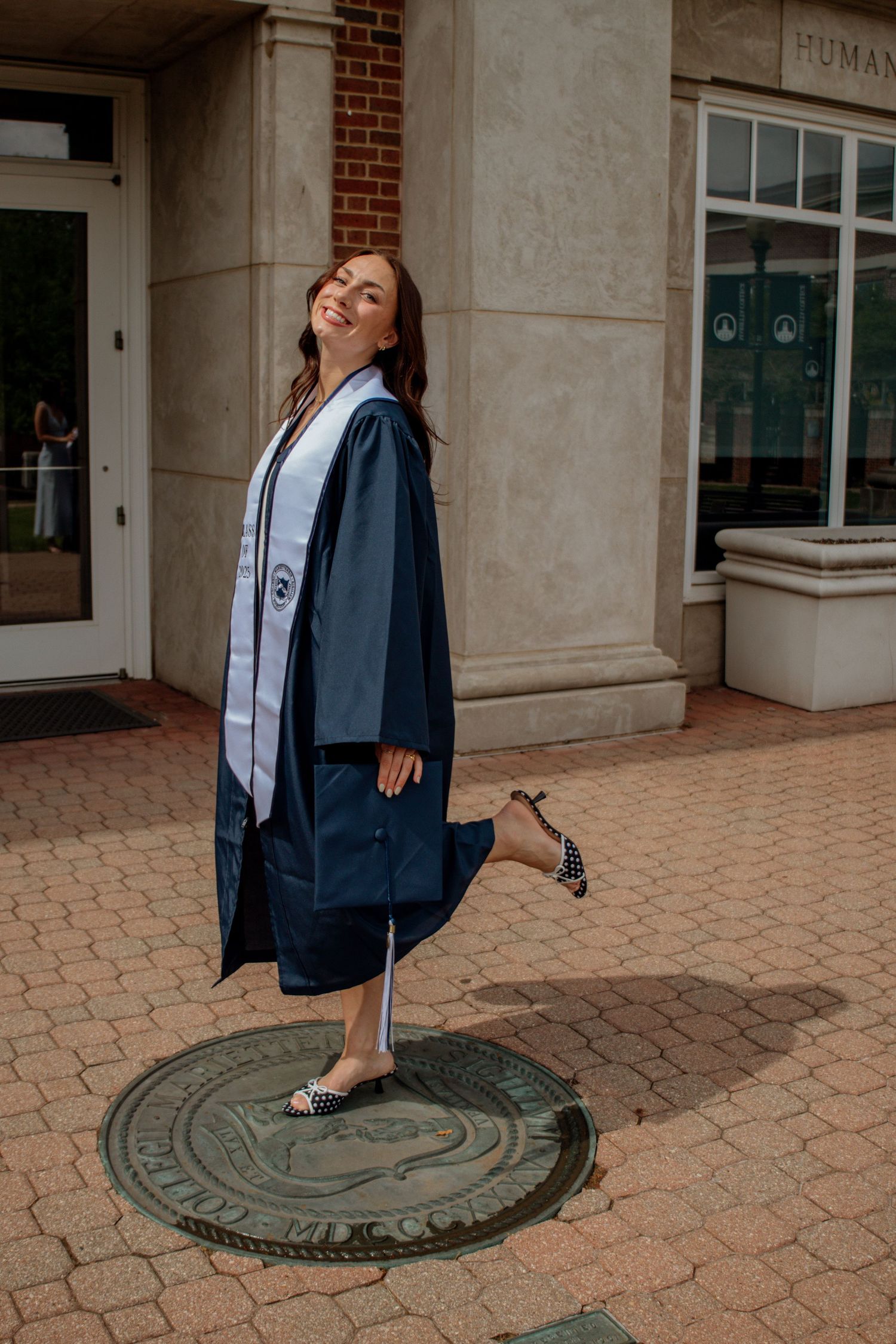 Smiling graduate in navy cap and gown poses playfully on university seal medallion outside Legacy Library.