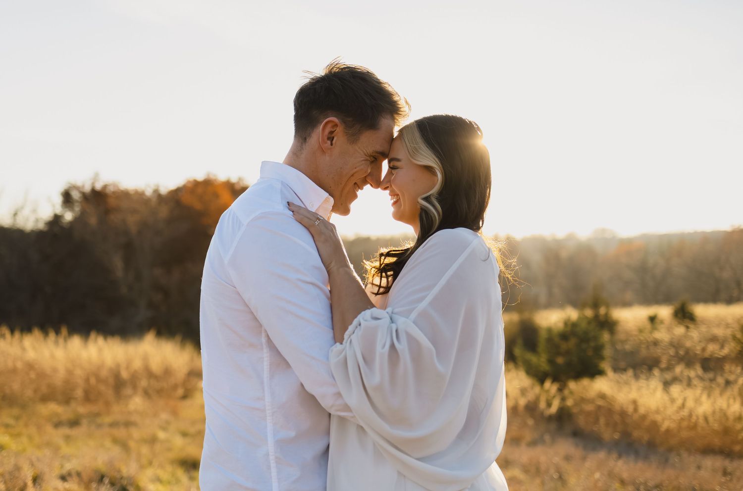 Couple embracing at golden hour in autumn field, foreheads touching romantically, wearing white outfits outdoors.