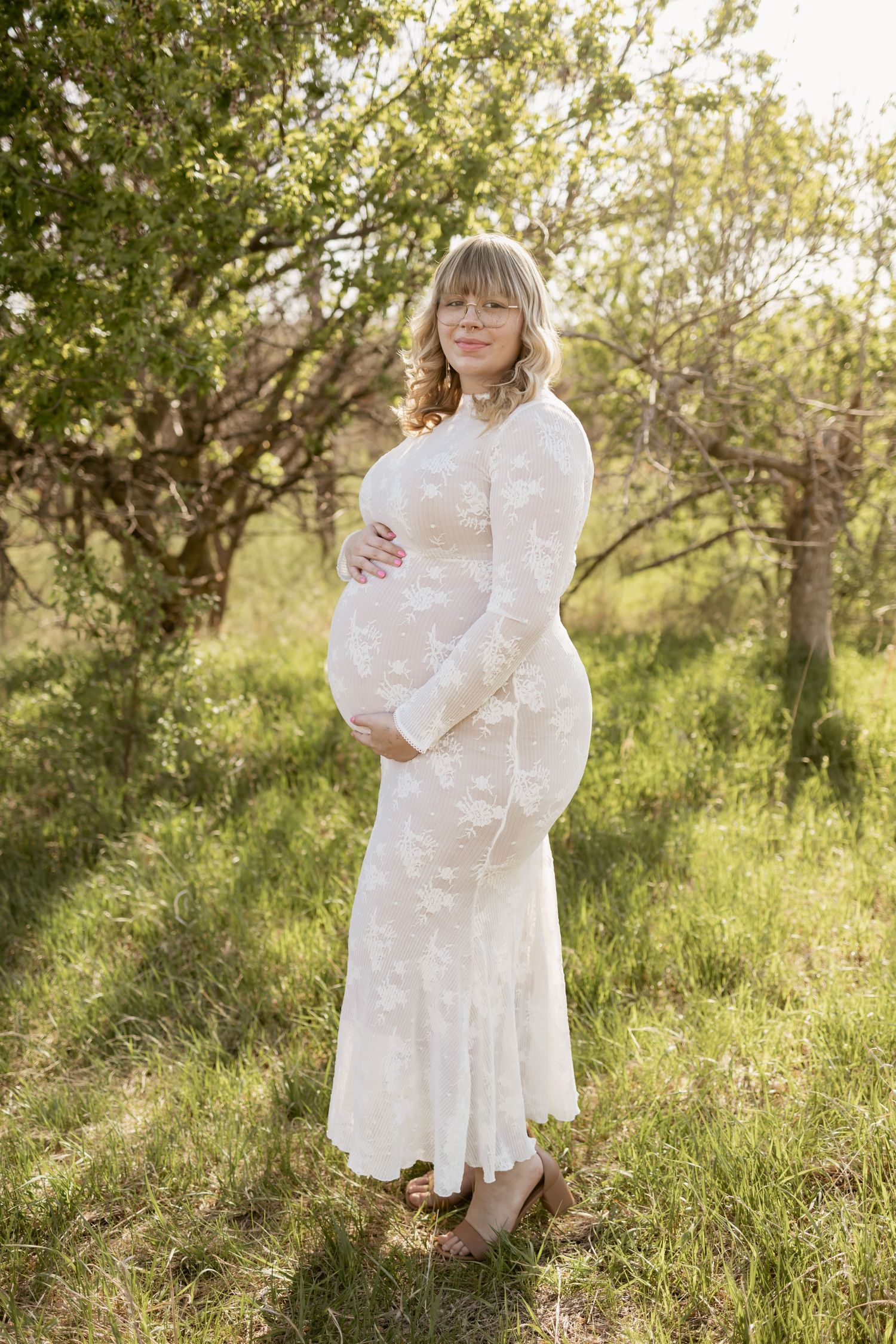 Pregnant woman in white lace dress holding baby bump outdoors among blooming trees in golden sunlight.