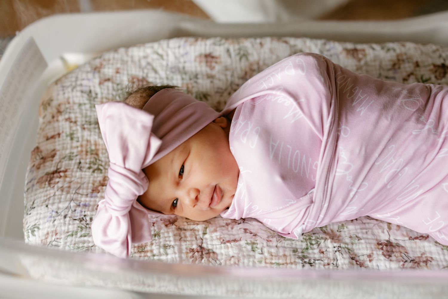 Newborn baby swaddled in pink blanket wearing a pink bow hat, sleeping peacefully in a white floral bassinet.