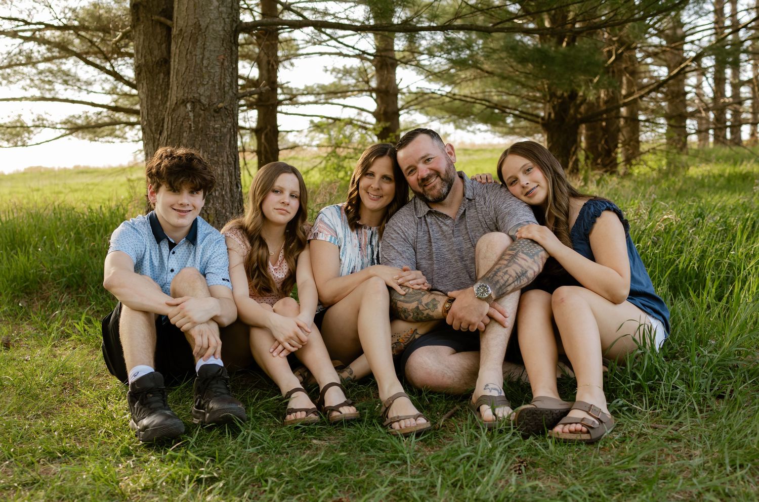 Family of five smiling together outdoors under pine trees on green grass in a warm summer portrait.