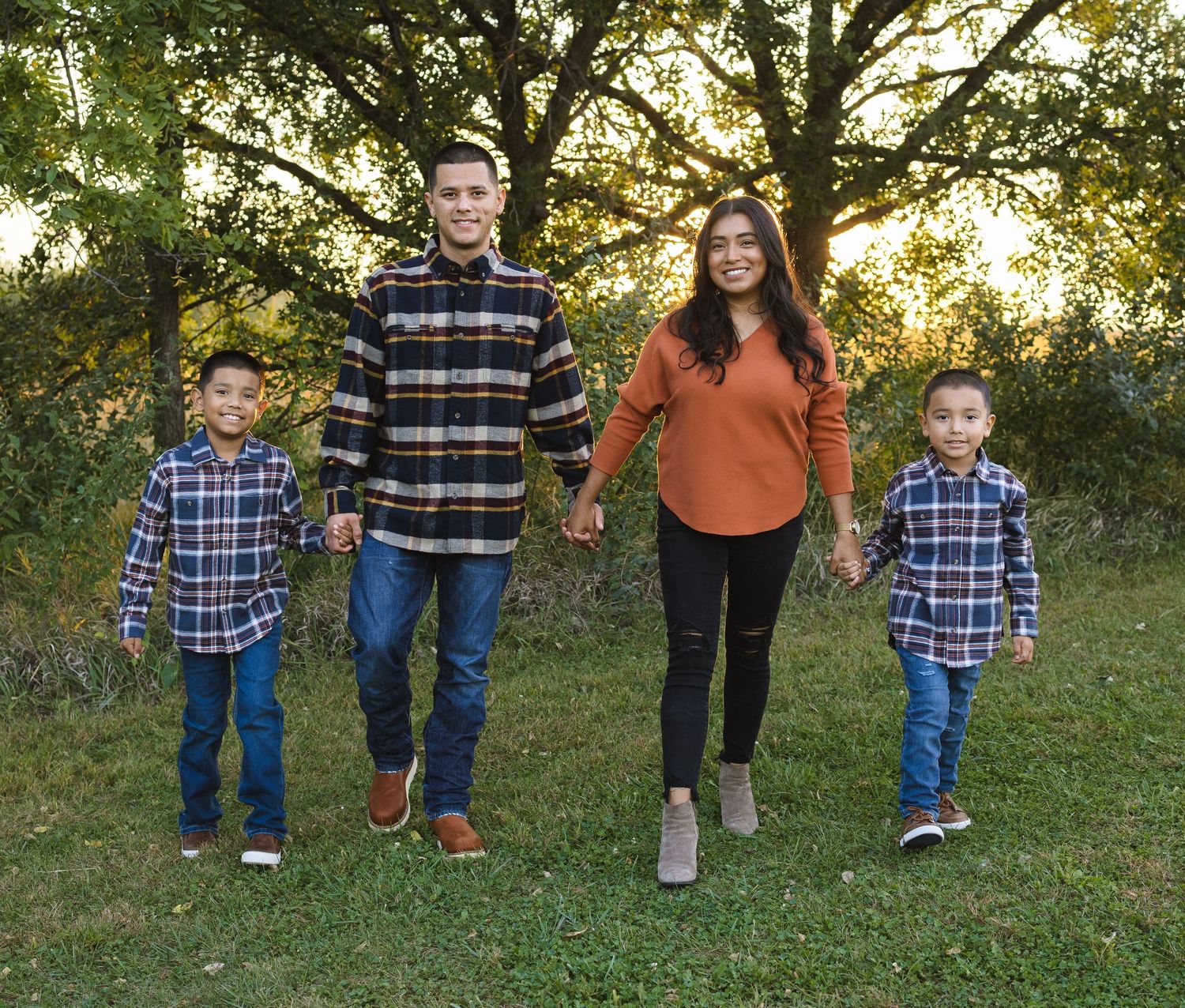 Family of four walking outdoors in coordinating flannel shirts, holding hands on a grassy lawn with trees in the background.
