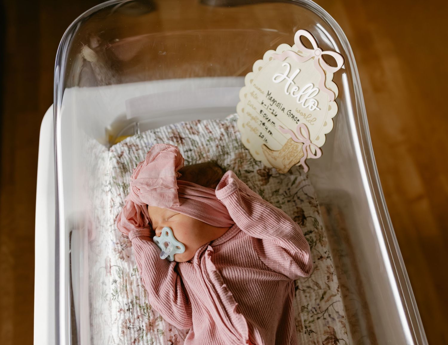 Newborn baby in pink outfit resting in hospital bassinet with a Hello name card.