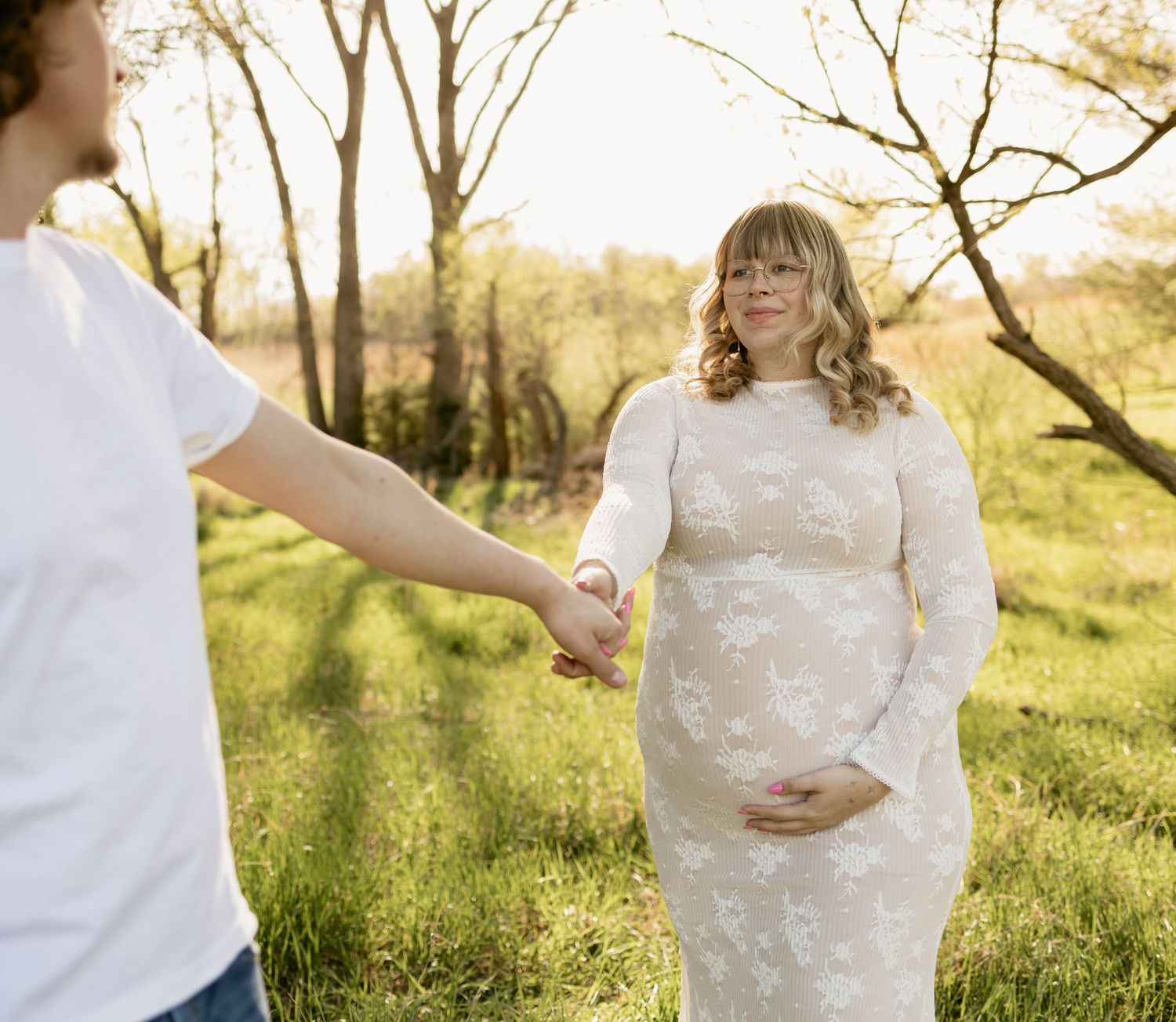 Pregnant woman in white lace dress holds hands with partner in sunny green field during maternity photoshoot.