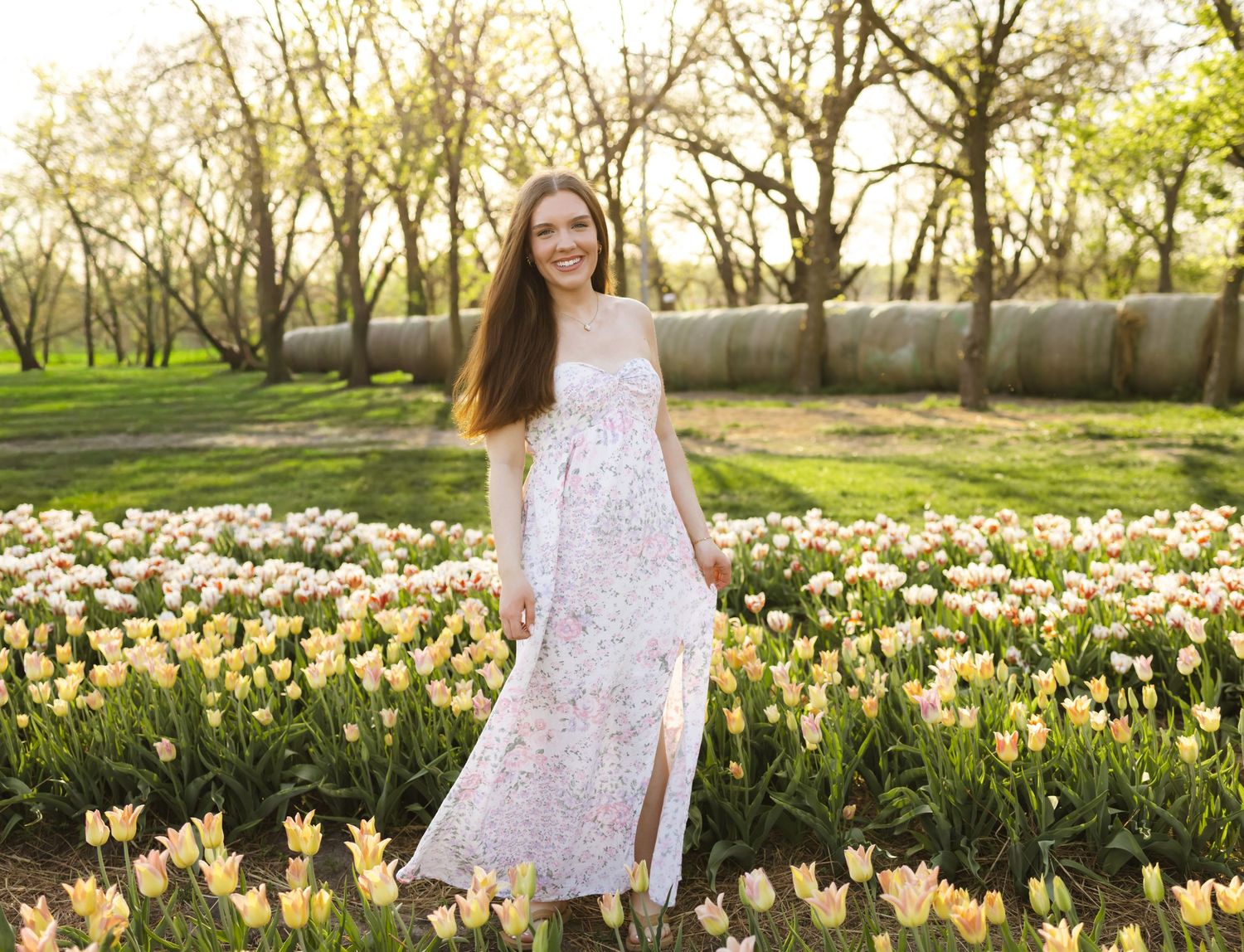 Young woman in floral maxi dress standing in colorful tulip garden with blooming trees in background.