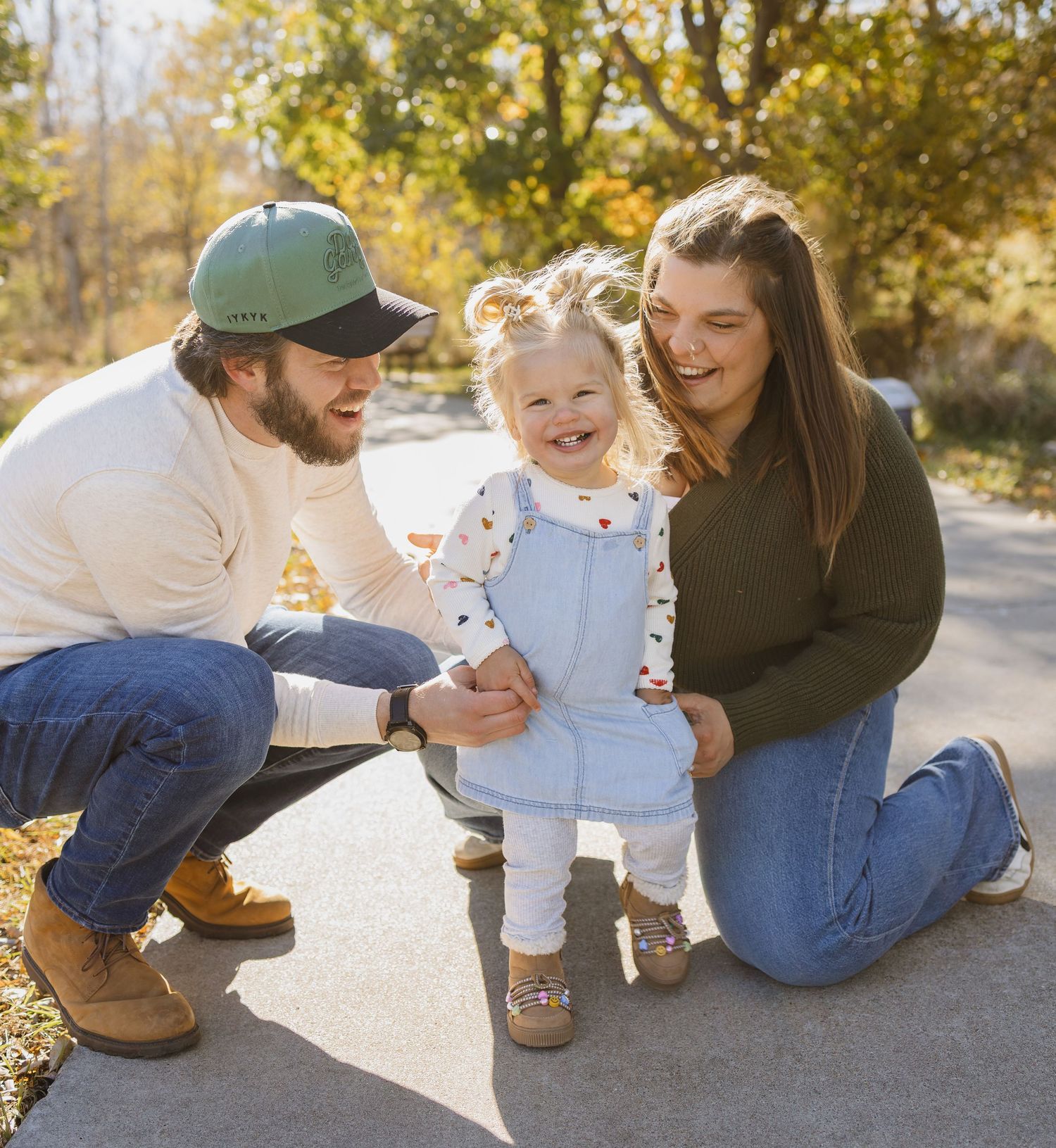 Happy family with toddler daughter smiling outdoors in autumn, parents crouching beside child on sunny day.