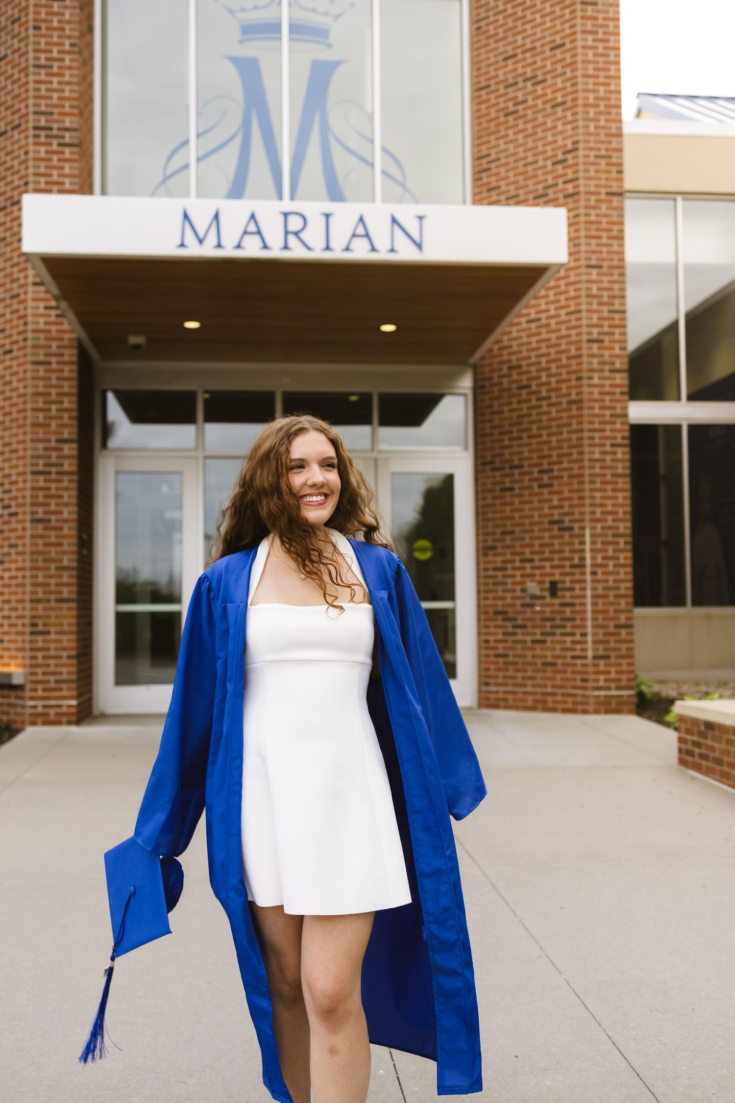 A smiling graduate in a blue cap and gown poses outside Marian University in a white dress.