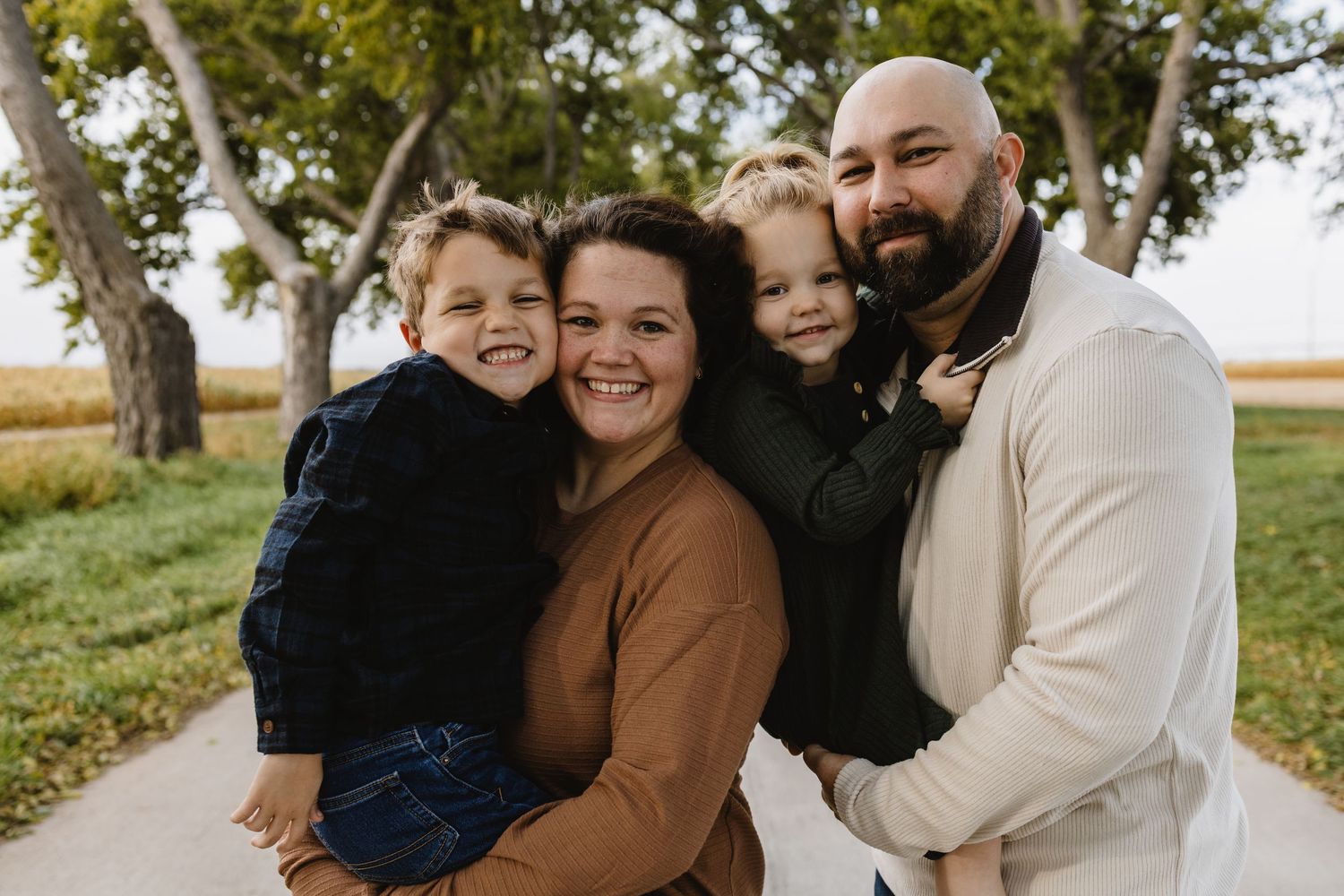 Happy family of four smiling outdoors, parents holding two young boys, trees and green landscape in background.