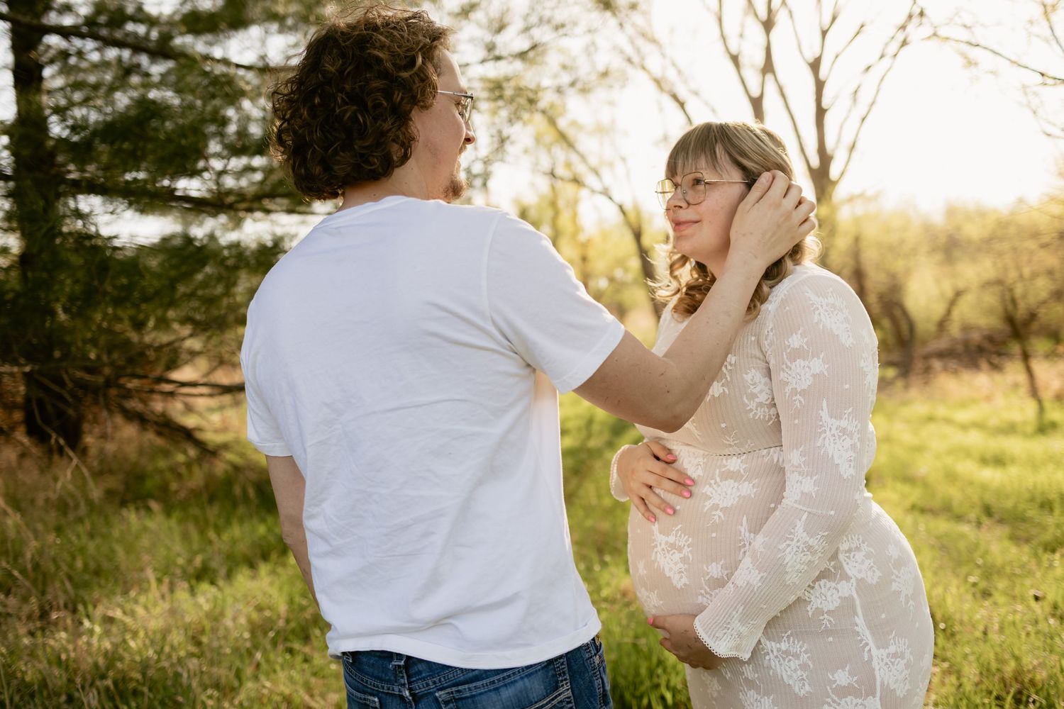 Couple sharing a tender moment outdoors during golden hour, pregnant woman smiling as man gently touches her hair.