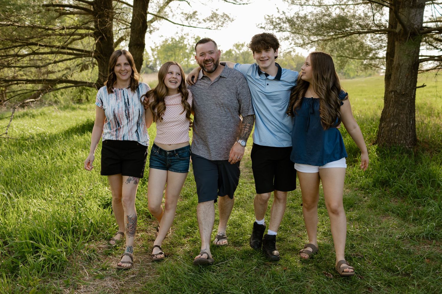 Happy family of five walking together outdoors on a grassy path surrounded by trees on a sunny day.