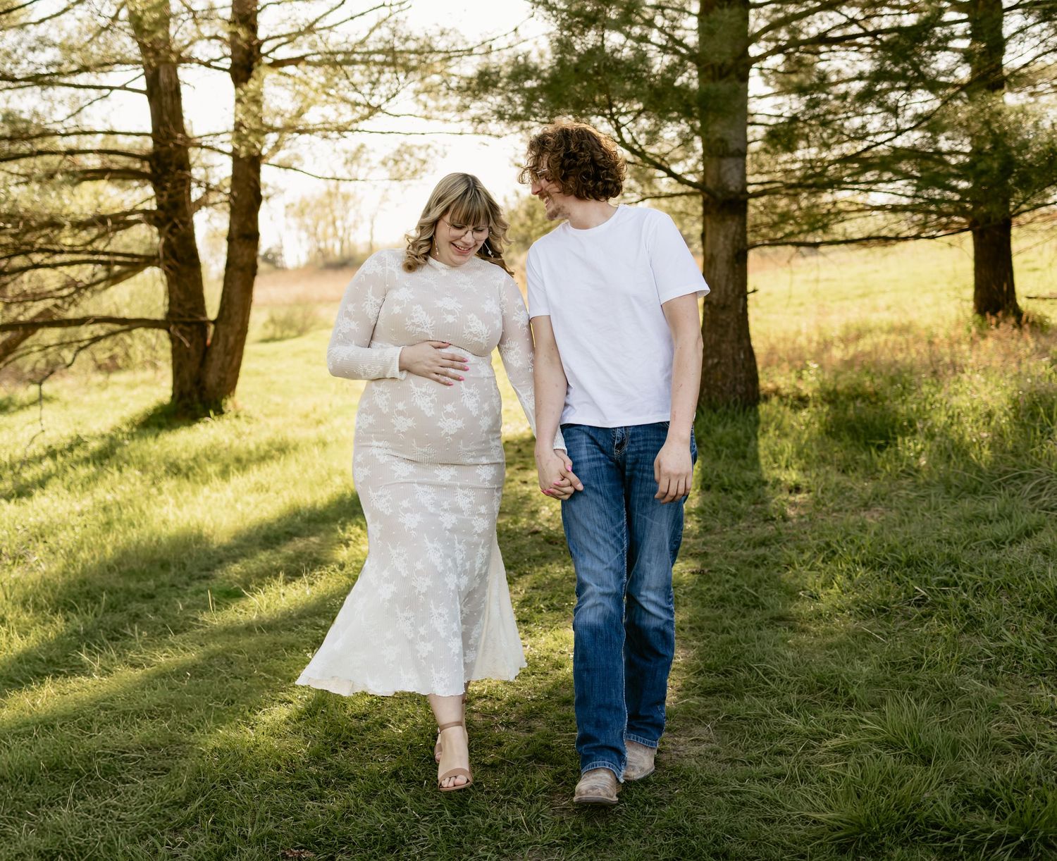Pregnant couple walking outdoors among trees, woman in white lace dress holding baby bump, man in white tee and jeans.