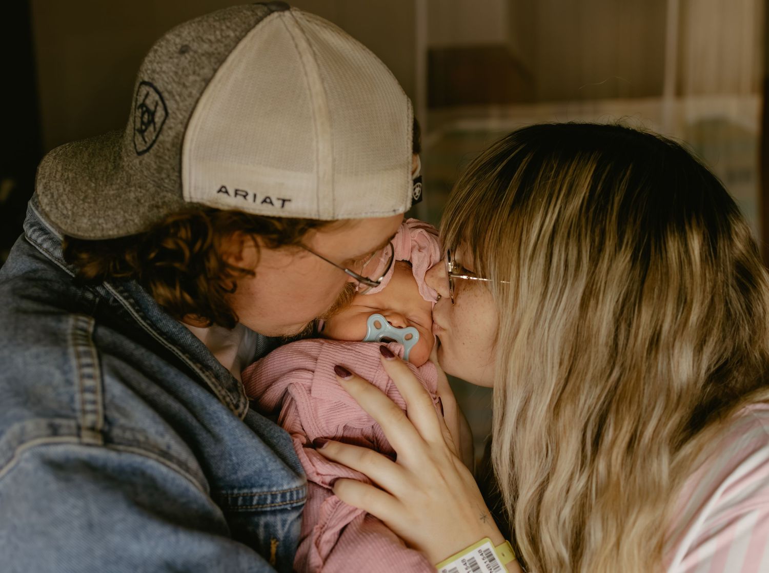 A couple shares a tender kiss over their newborn baby wrapped in a pink blanket.