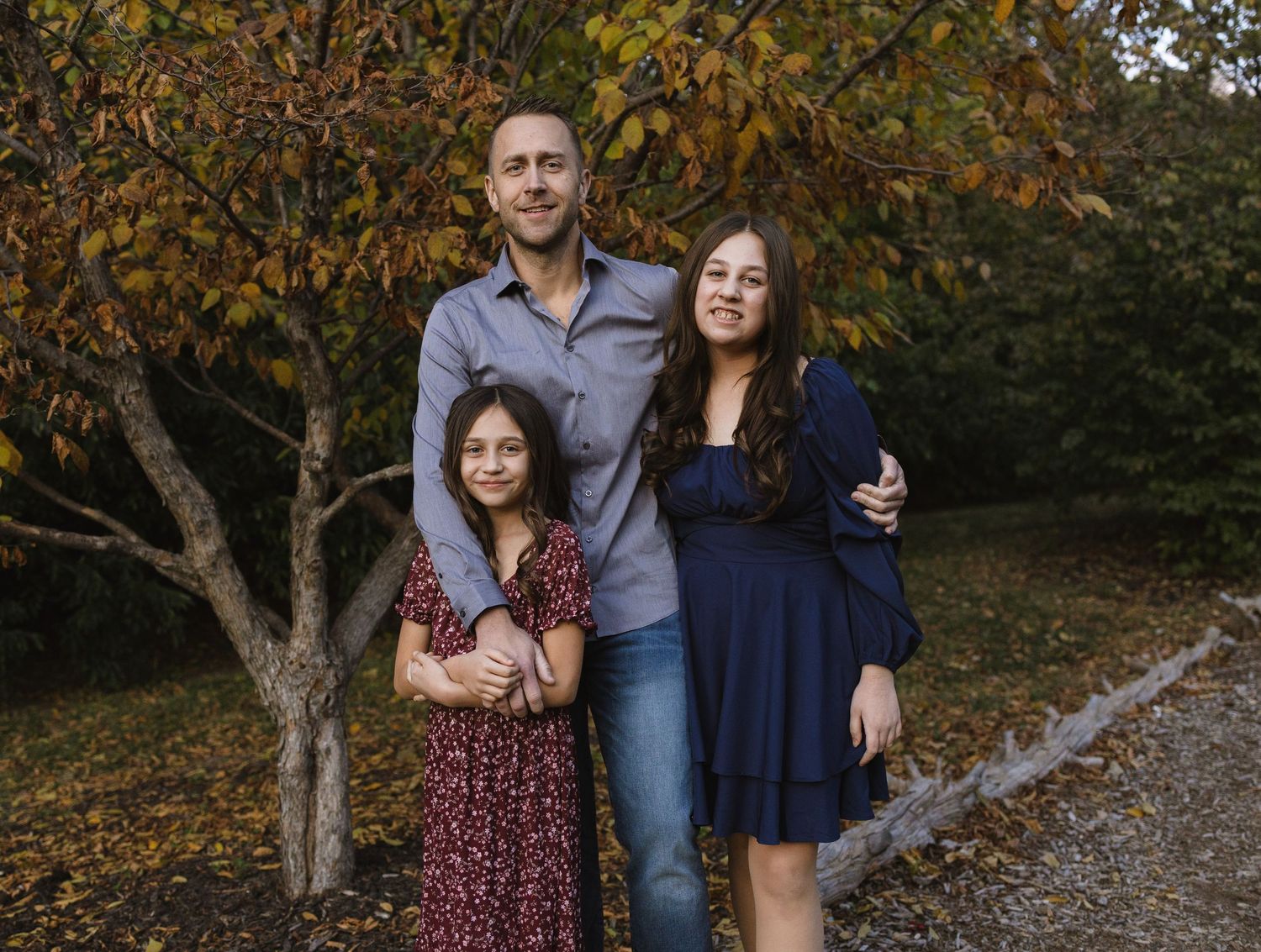 A man and two girls pose together outdoors among autumn trees on a gravel path for a family portrait.