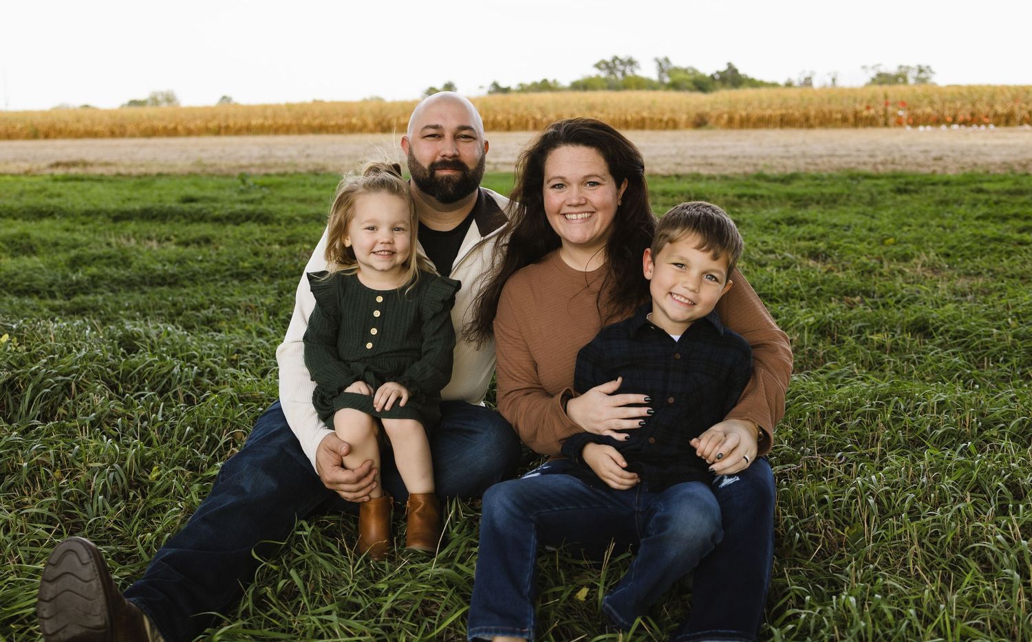 Happy family of four posing together in a green field with golden cornfields in the background.
