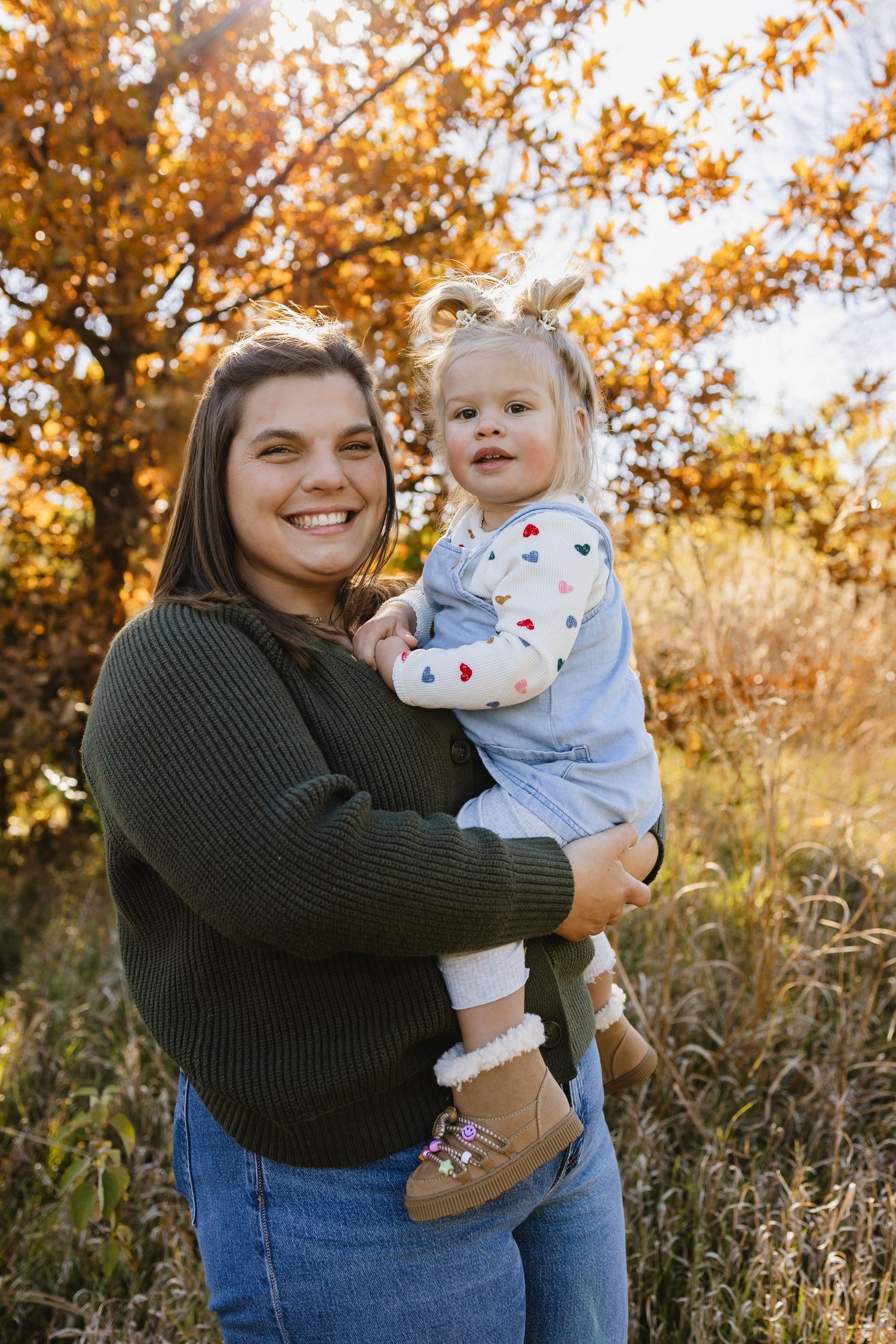 A smiling woman in a green sweater holds a toddler girl outdoors among autumn foliage in golden light.