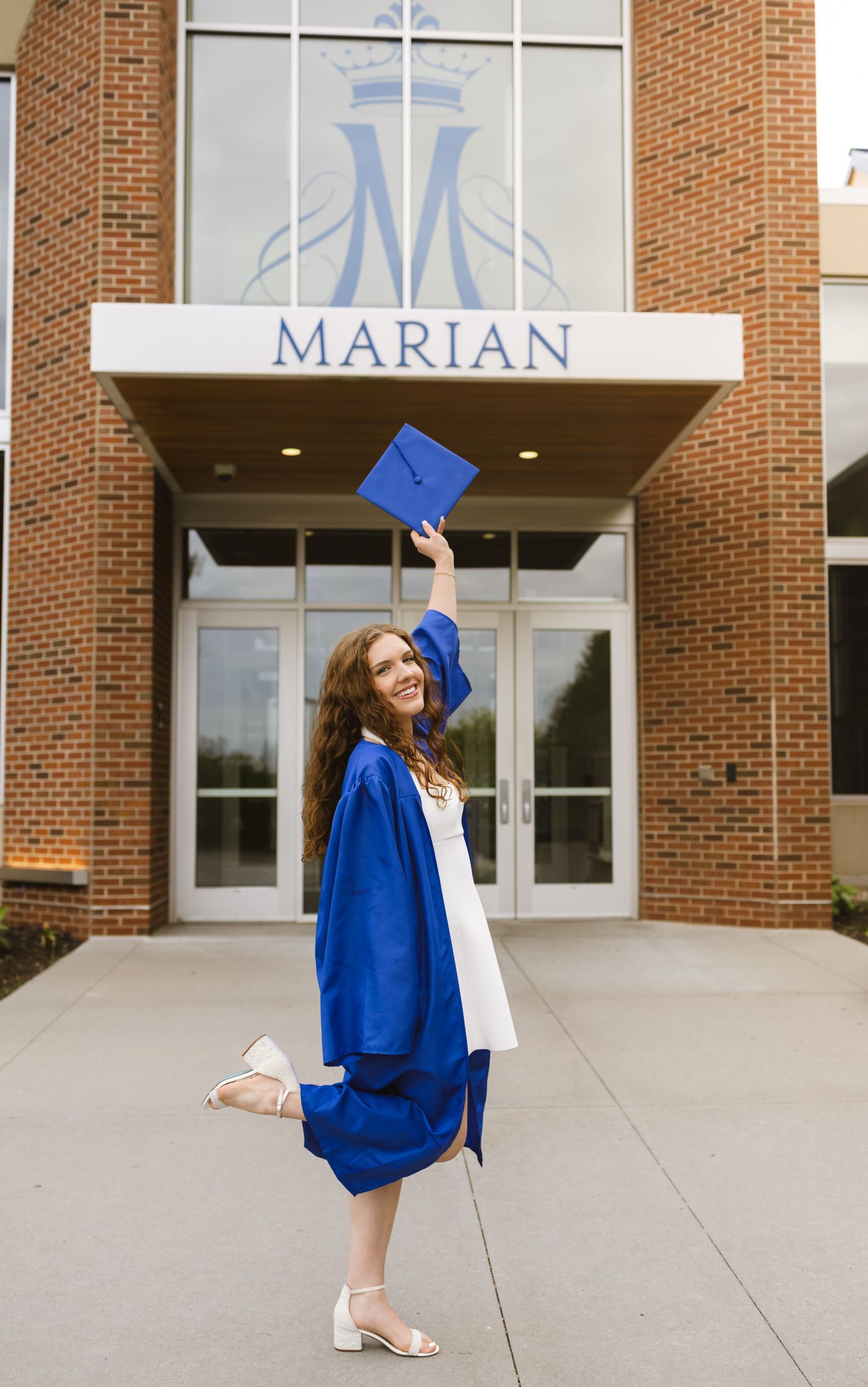 A young woman in blue graduation gown tosses her cap in front of Marian school building entrance.