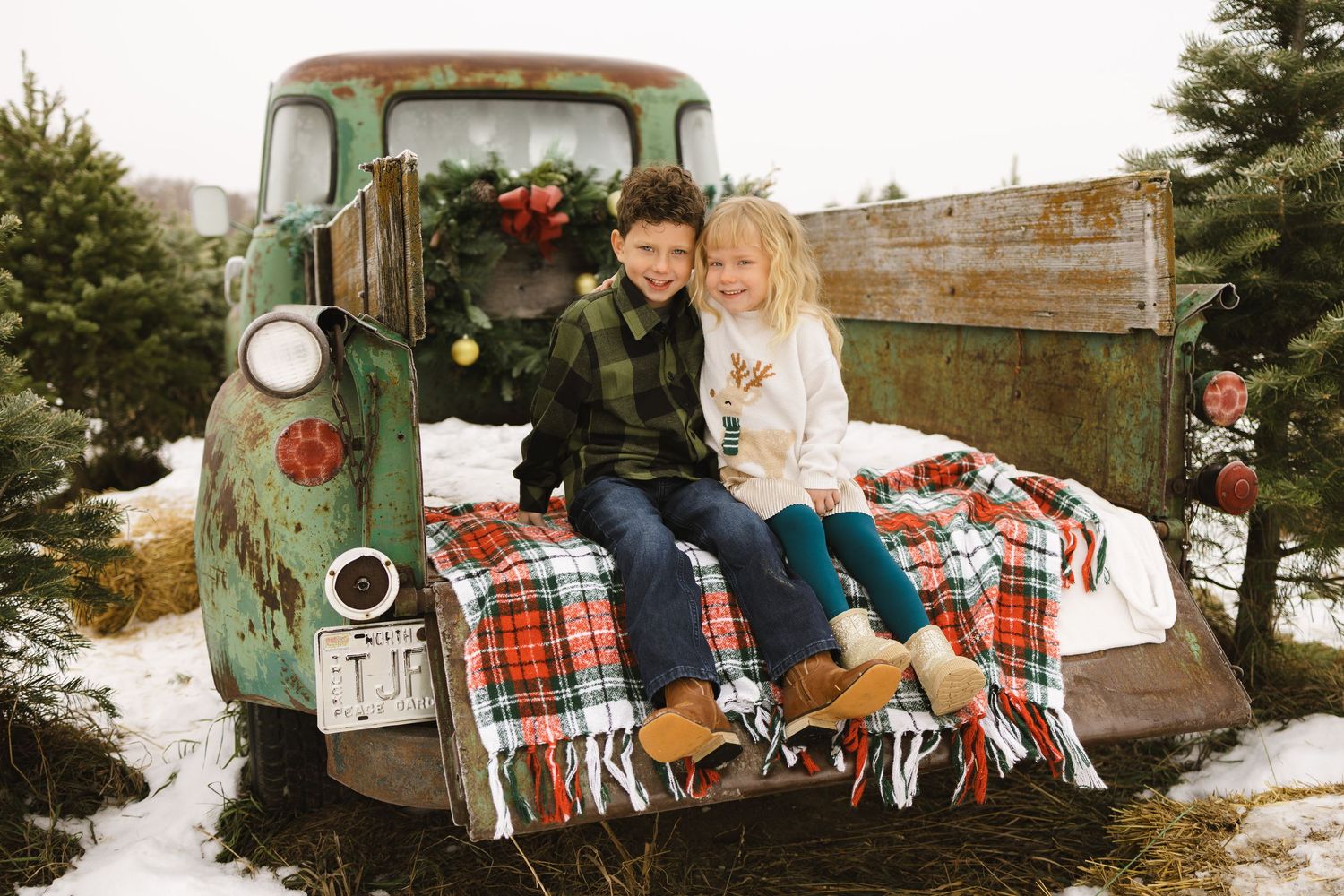 Two children sit on a vintage green truck bed with plaid blanket at a snowy Christmas tree farm.