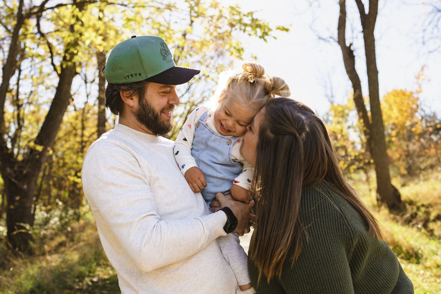 Happy young family with bearded dad in green cap and mom kissing toddler outdoors in sunny autumn forest.
