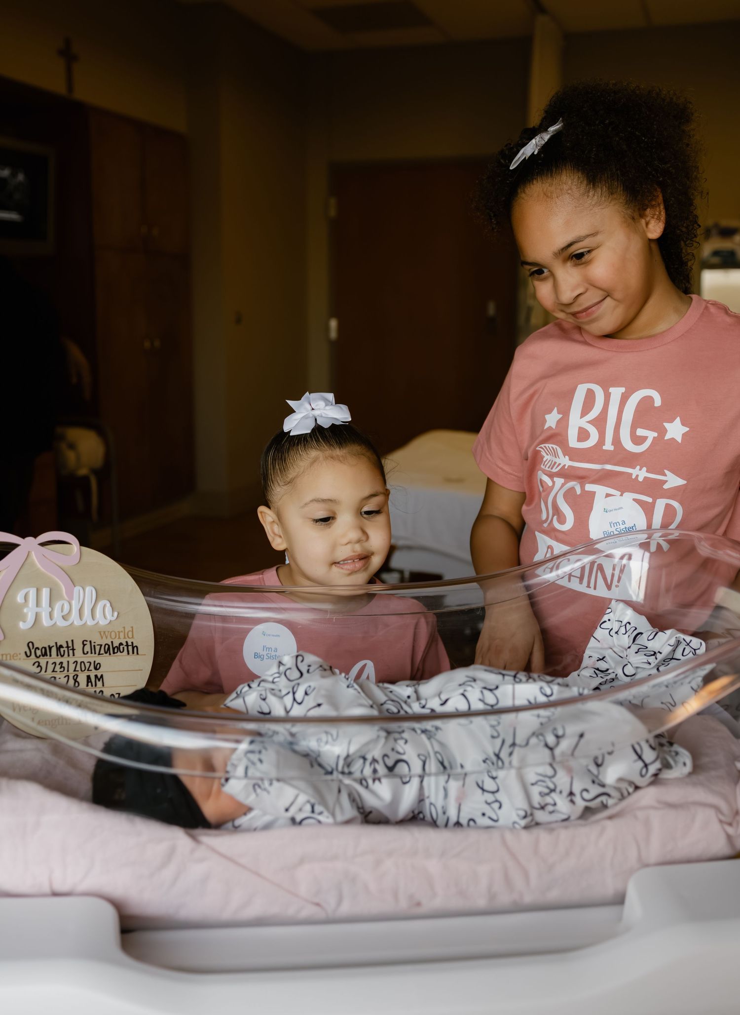 Two sisters admire their newborn baby sibling in a hospital bassinet, with a Hello name tag nearby.