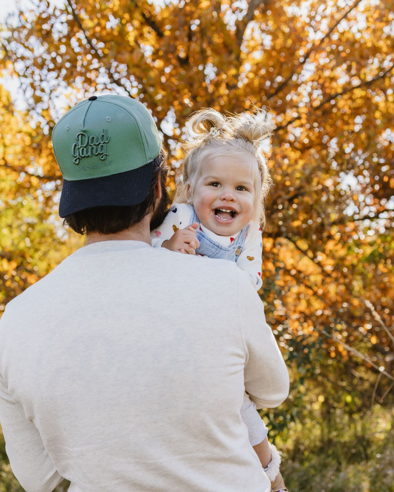 A father in a green cap lifts his laughing toddler girl against a colorful autumn tree backdrop.